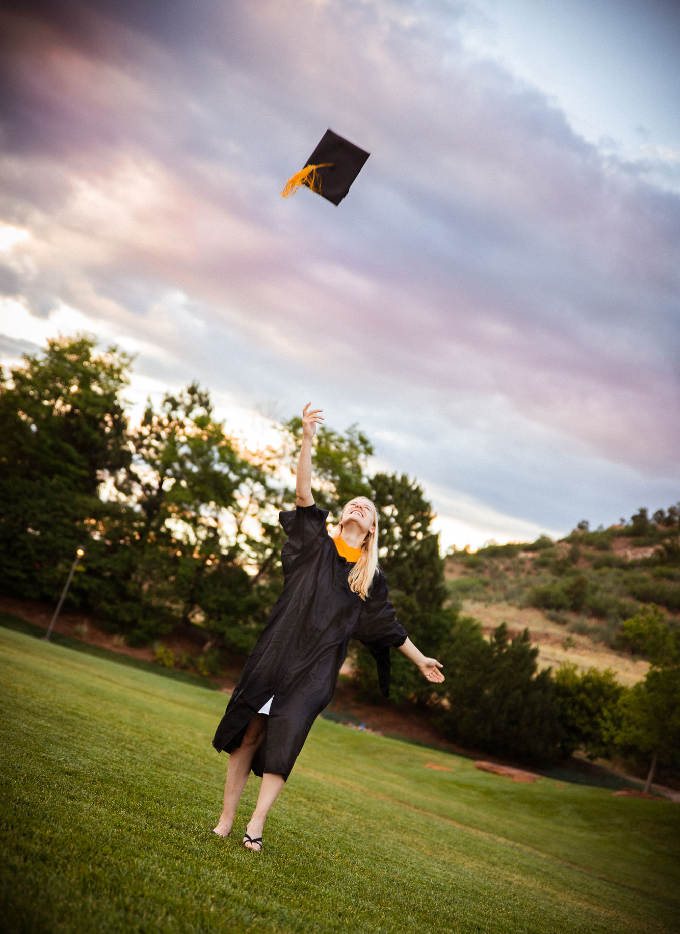 A graduate in black cap and gown joyfully tosses her mortarboard into the sky on a green lawn at sunset.