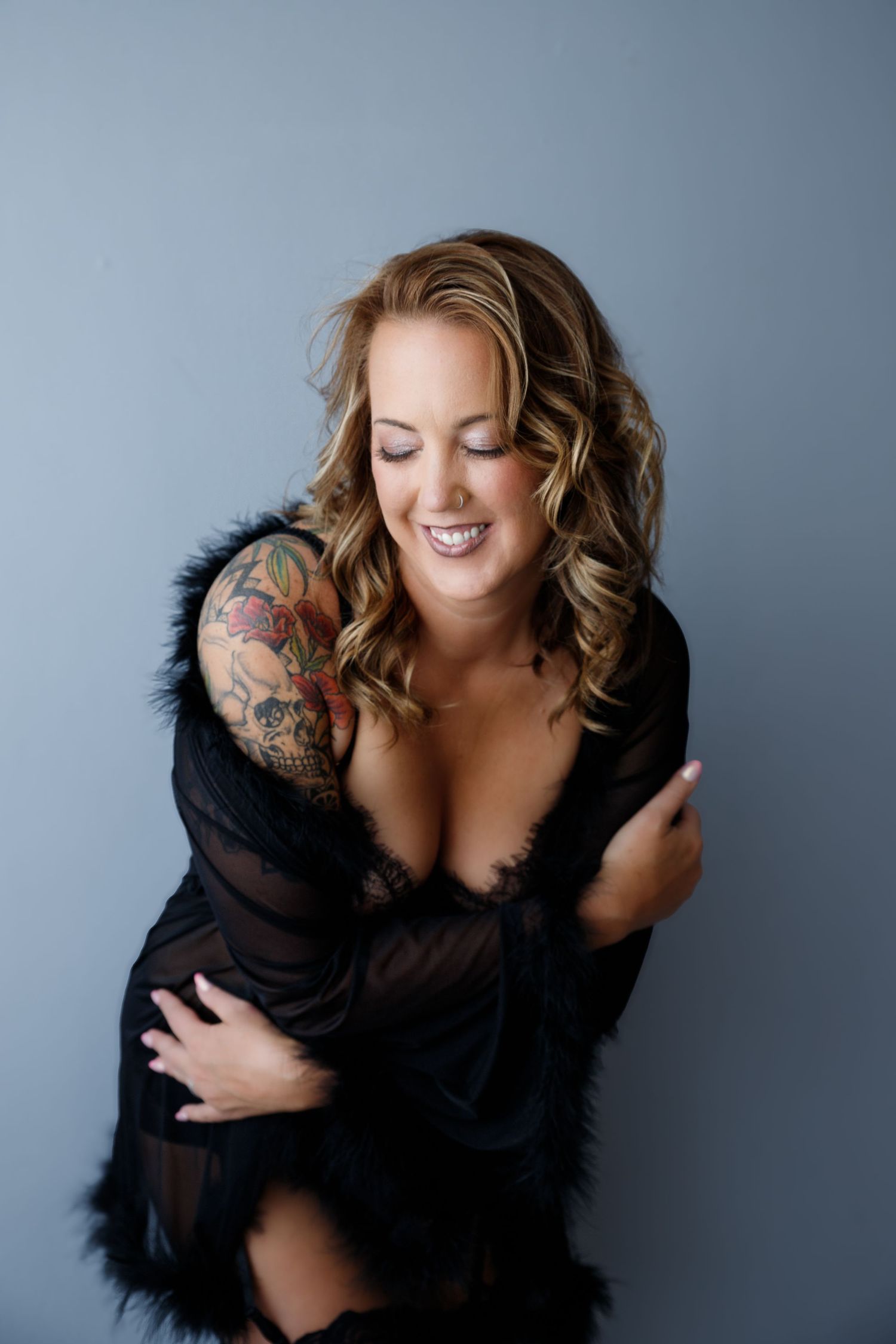 Smiling blonde woman with tattoos wearing black sheer robe with fur trim posing against gray background.