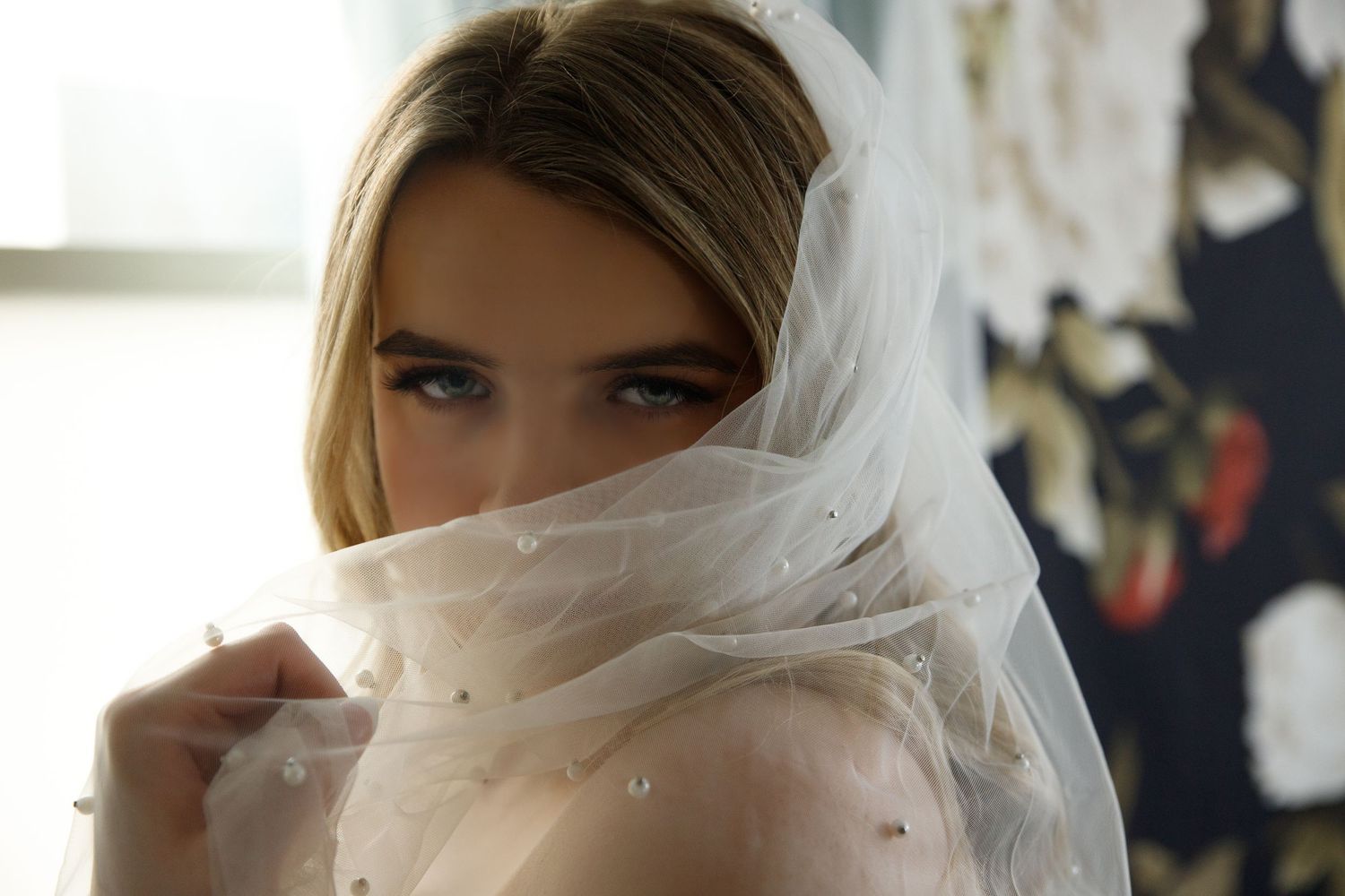 Young blonde woman holding a pearl-embellished white bridal veil over her face, with soft natural lighting.