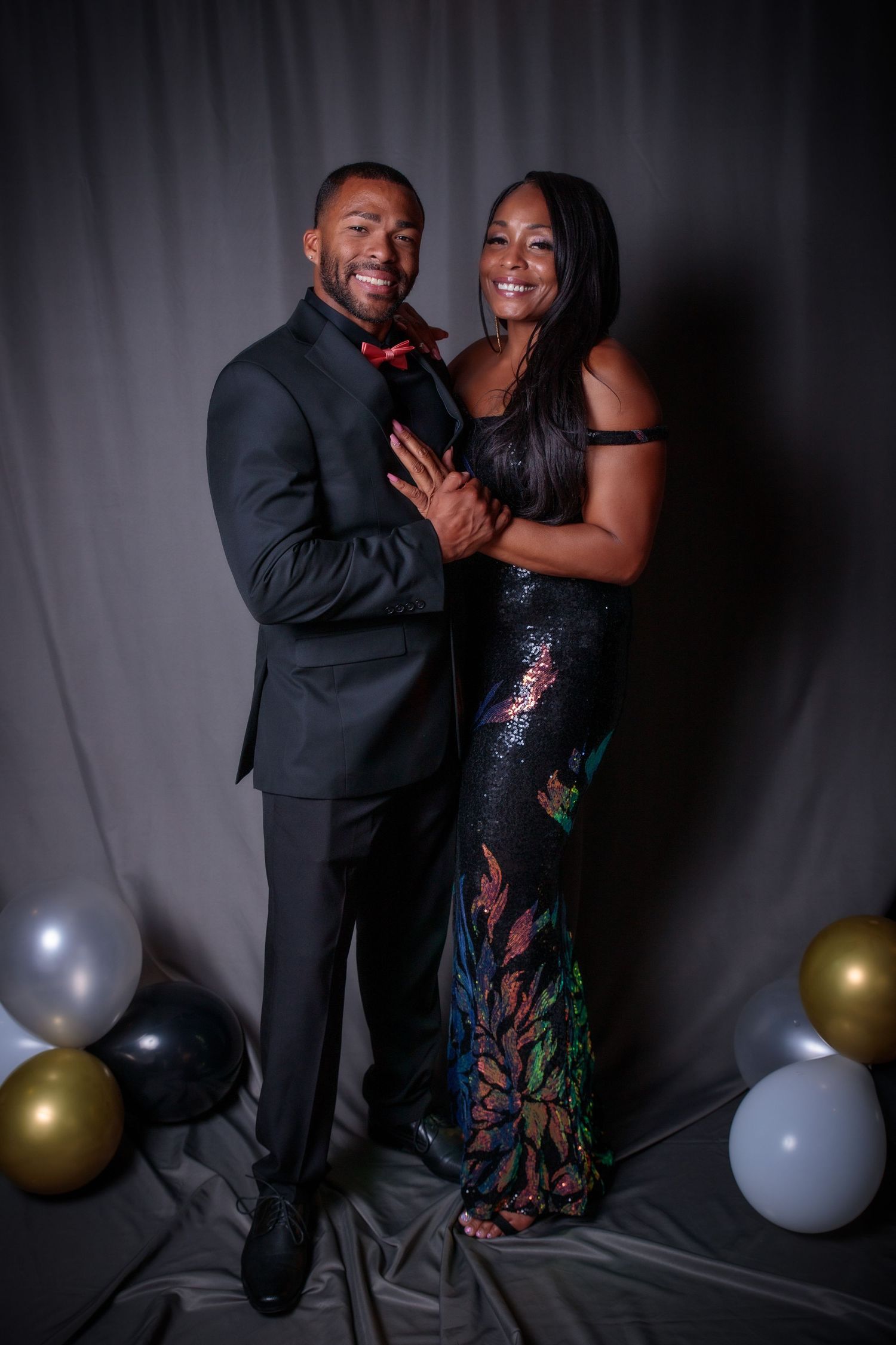 Smiling couple at formal event, man in black tuxedo with red bow tie and woman in multicolor sequin gown.