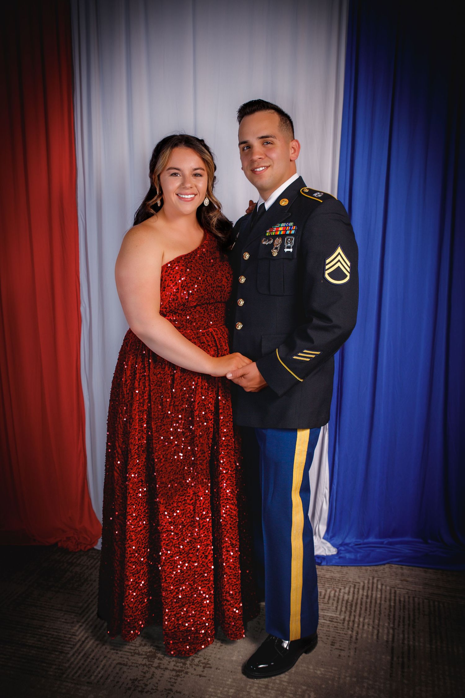 Couple posing at military ball, woman in red sequin gown and man in Army dress uniform with medals.