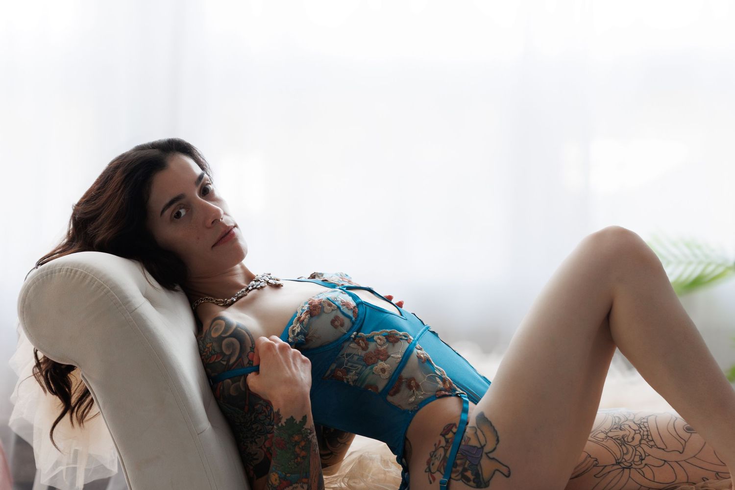 Tattooed woman in a teal bodysuit reclining on a white chaise lounge in a bright, softly lit boudoir photography studio in Colorado Springs.