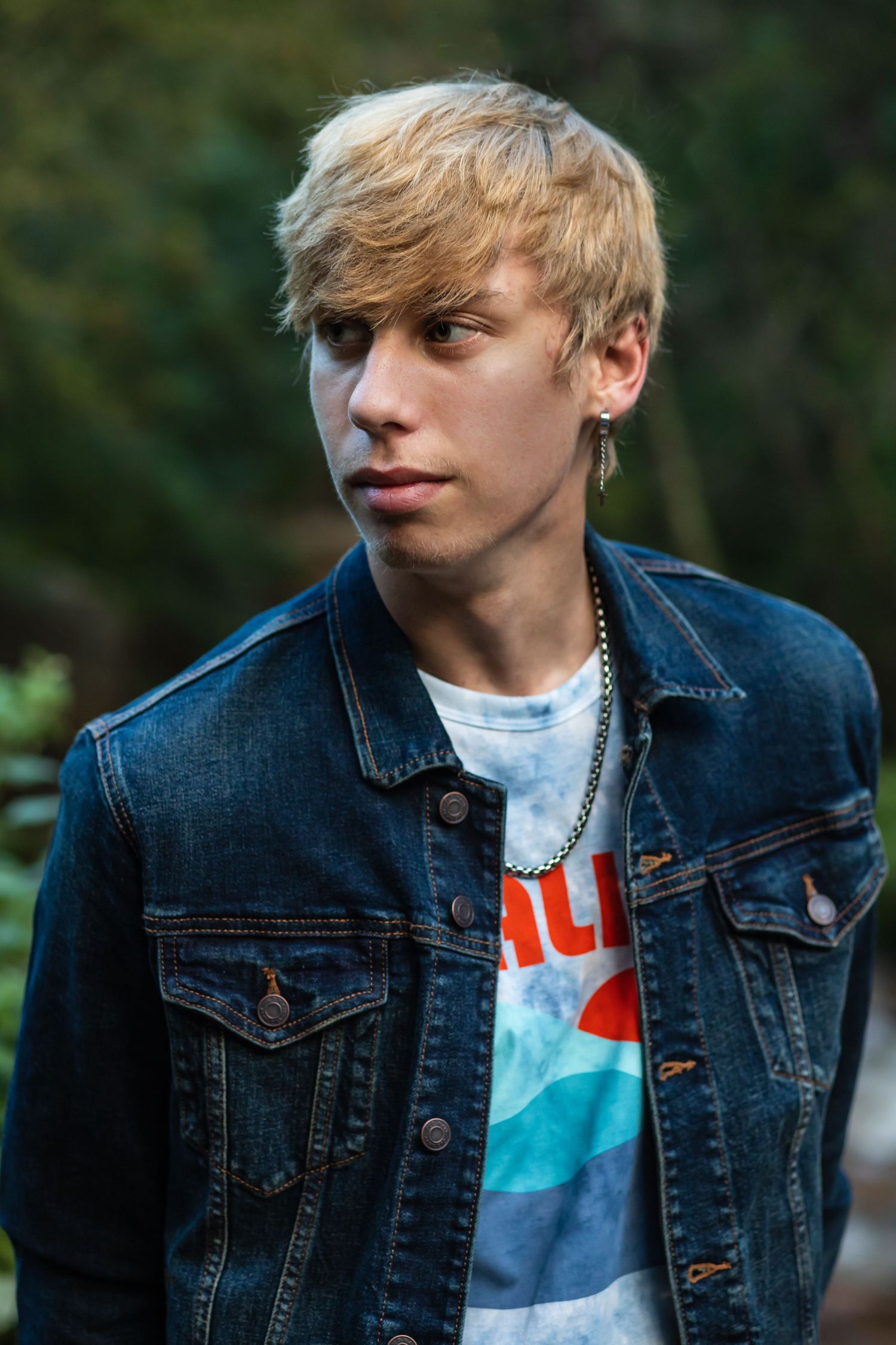 Young blonde teen in denim jacket and graphic tee with chain necklace poses outdoors near green foliage.