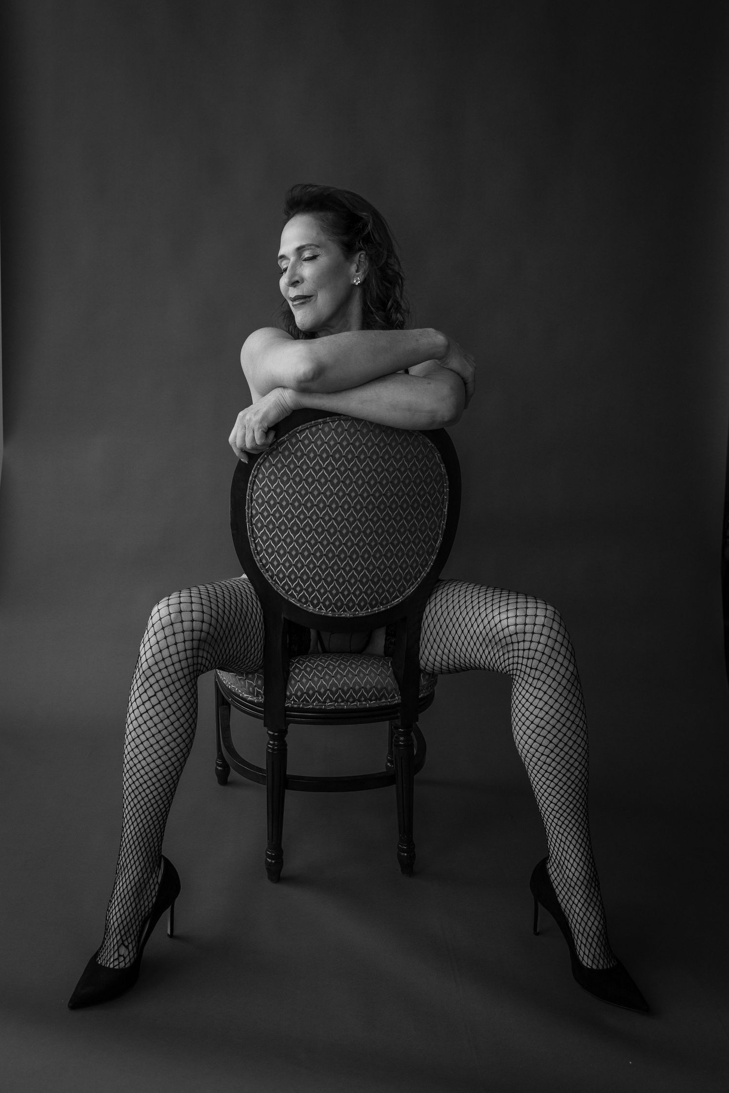 Black-and-white boudoir portrait of a woman posing on a vintage chair in stockings and heels, by Nicole Fey Photography in Colorado Springs
