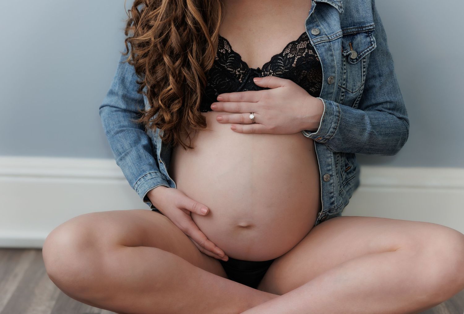Pregnant woman in denim jacket and black bra sitting cross-legged, cradling her baby bump with both hands.