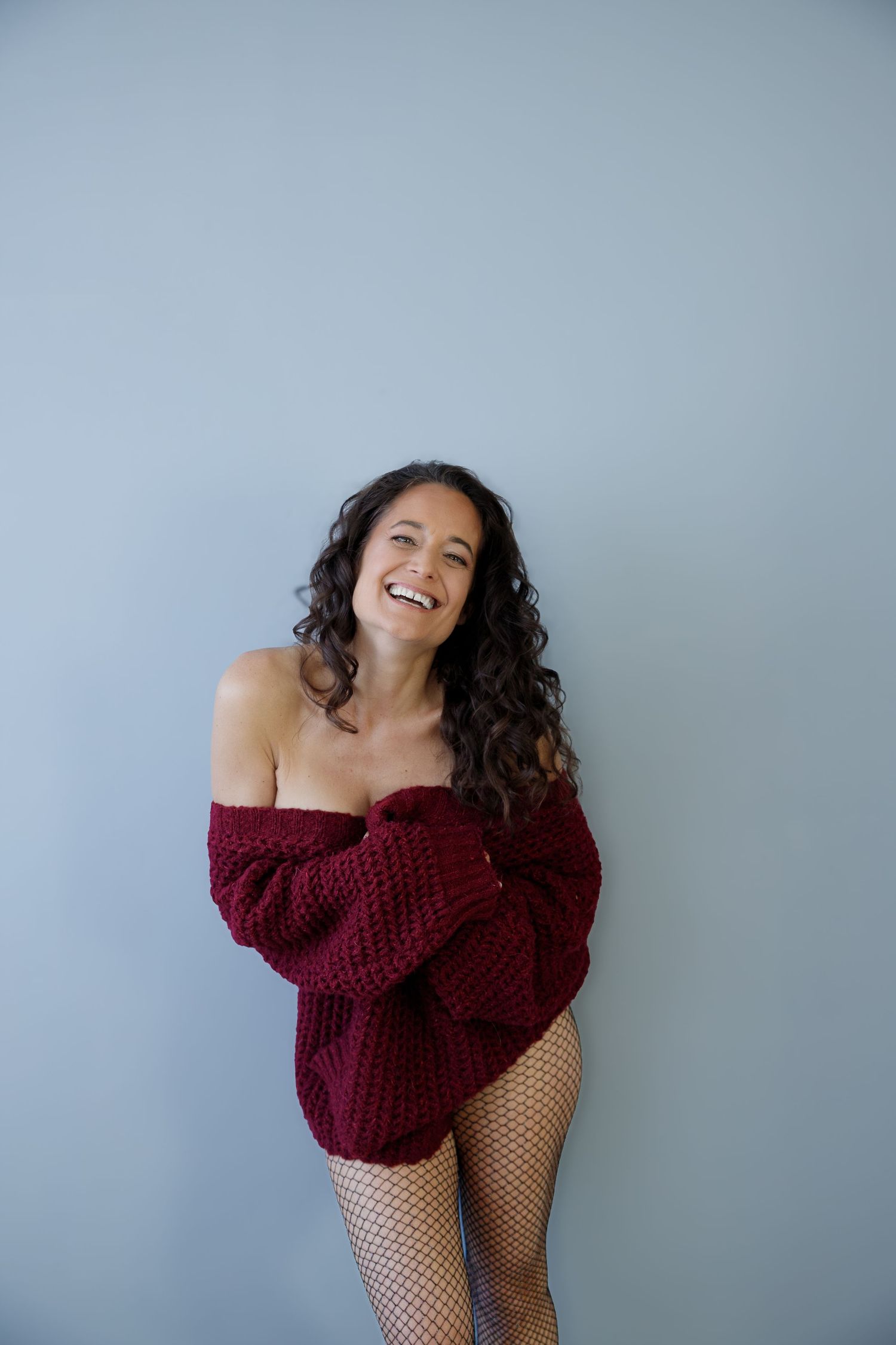 Laughing woman in burgundy off-shoulder knit sweater and fishnet tights posing against gray wall.