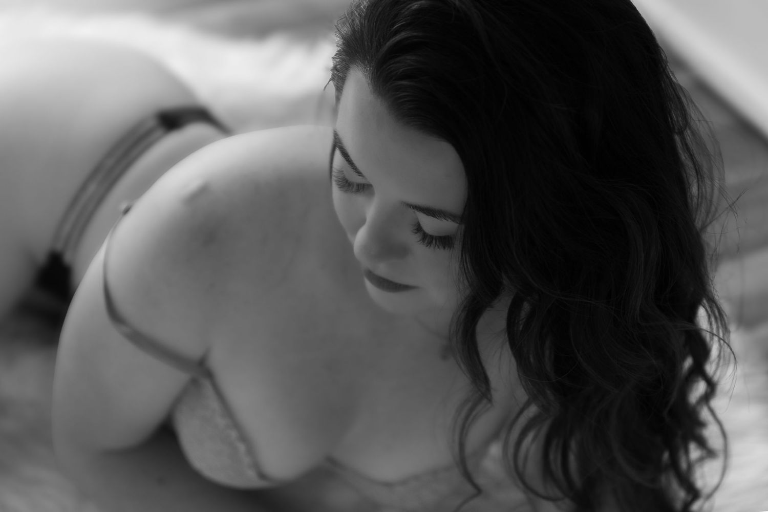Black and white boudoir photo of a curvy woman with dark curly hair on a bed.