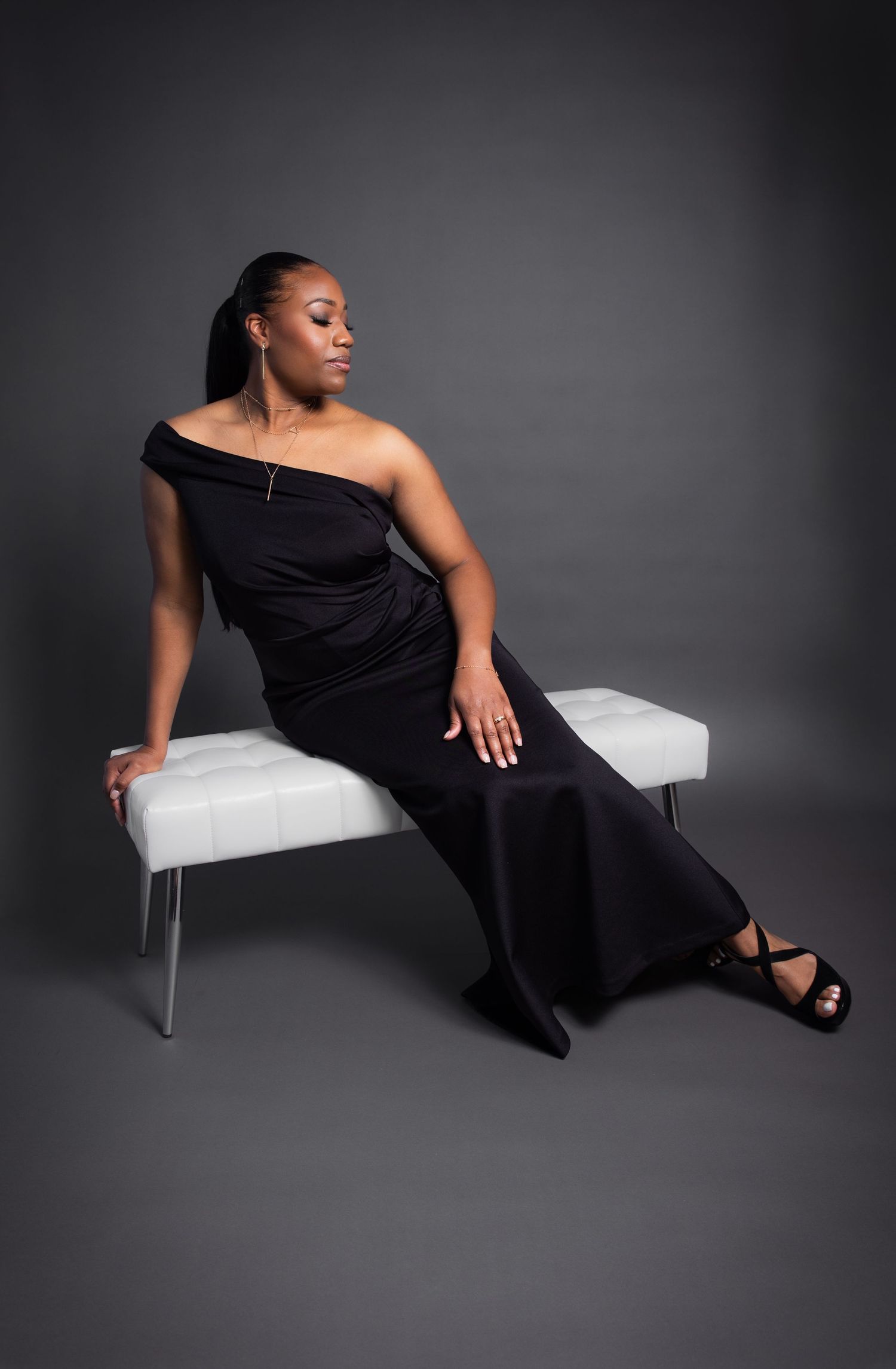 Woman in black off-shoulder gown seated gracefully on white bench.