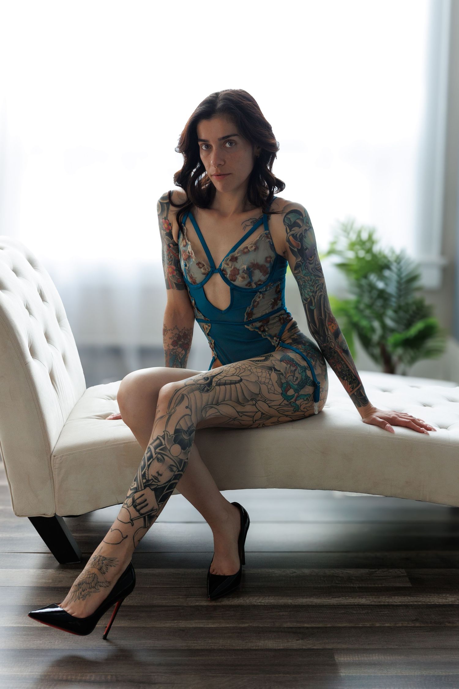 Tattooed woman in teal bodysuit sitting on white chaise lounge wearing black heels in bright modern boudoir studio in Colorado Springs.