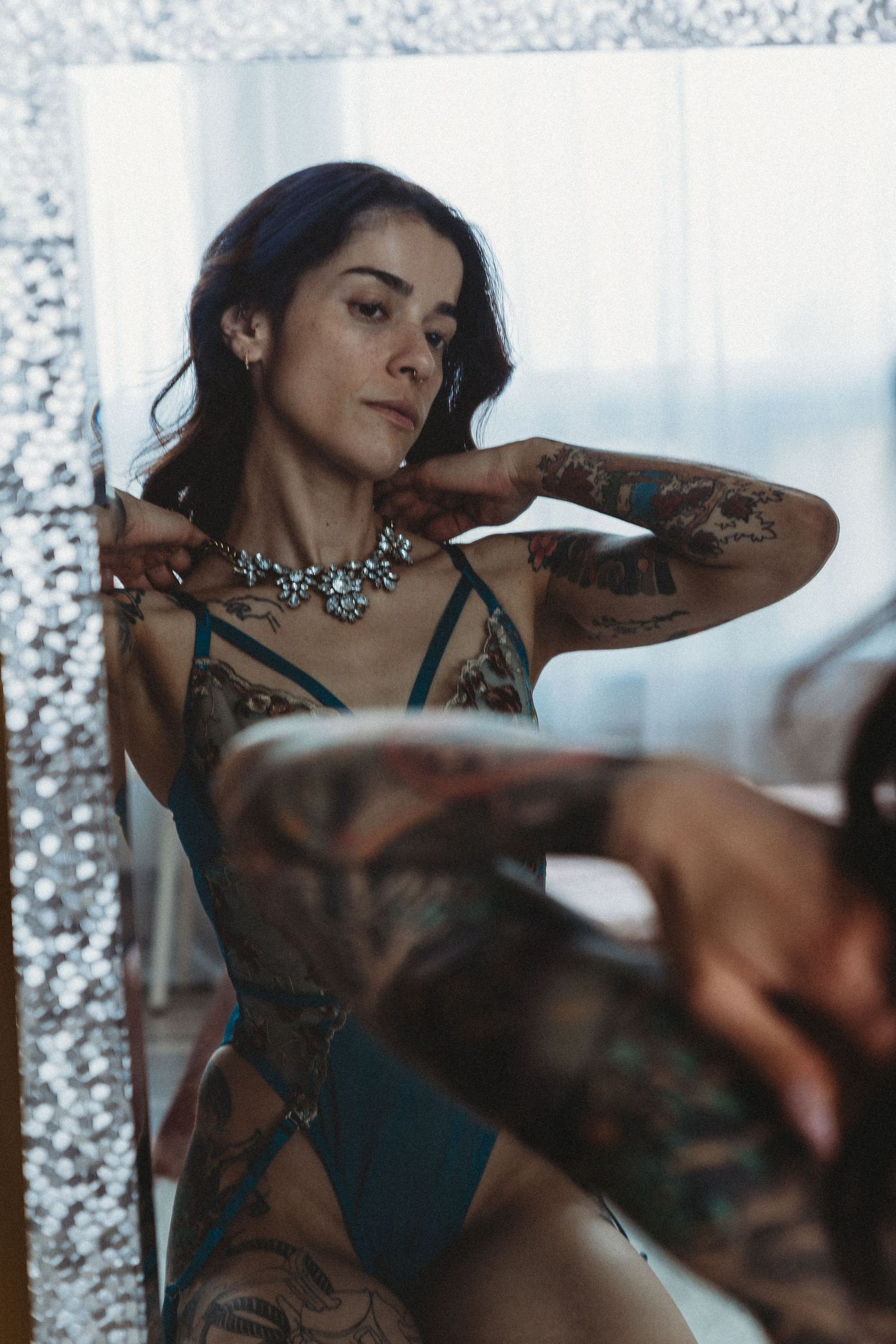 Tattooed woman in teal lingerie and statement necklace posing by a mirror in soft natural light.