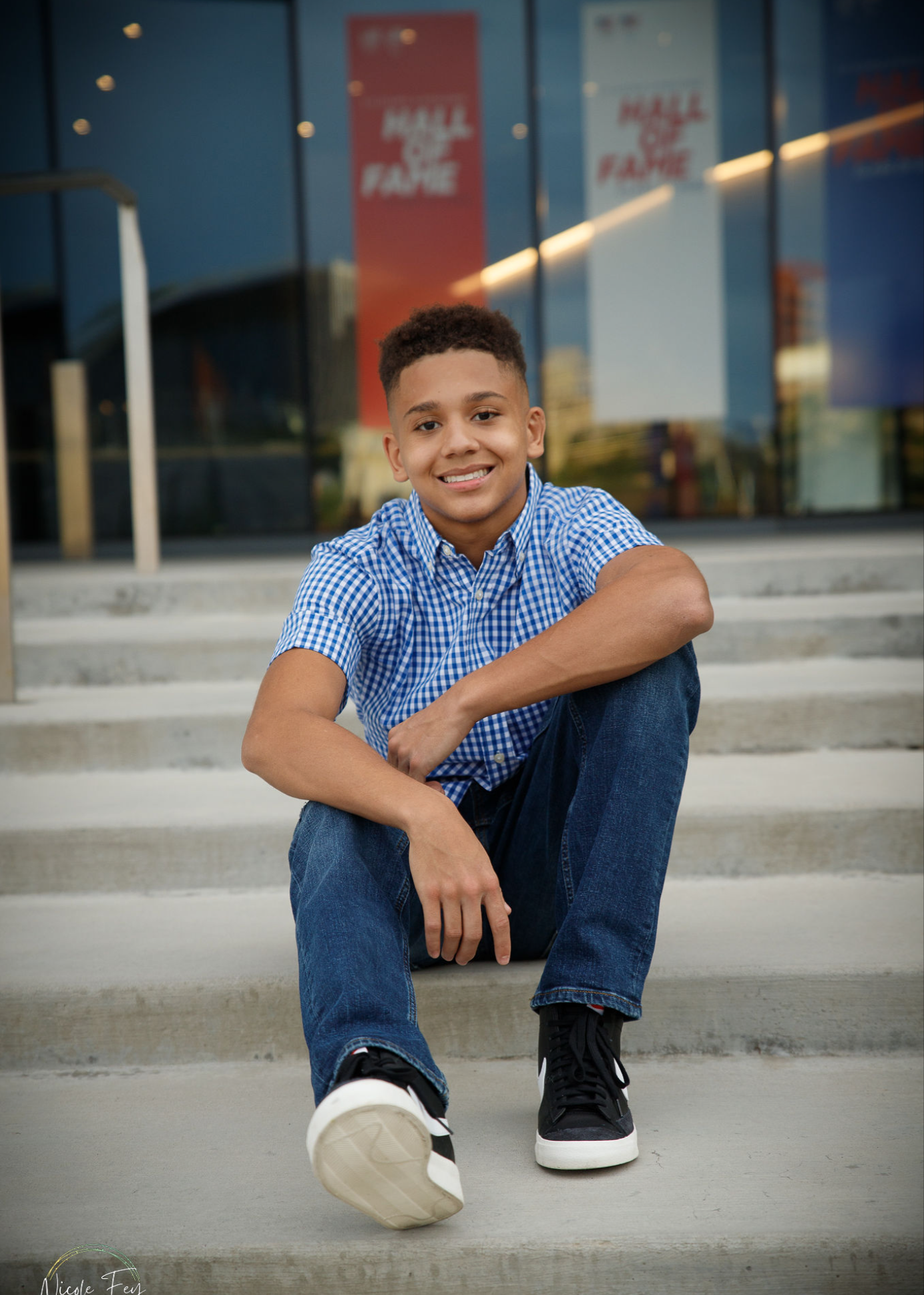 Smiling teenage boy in blue checkered shirt sitting on steps outside Hall of Fame building.