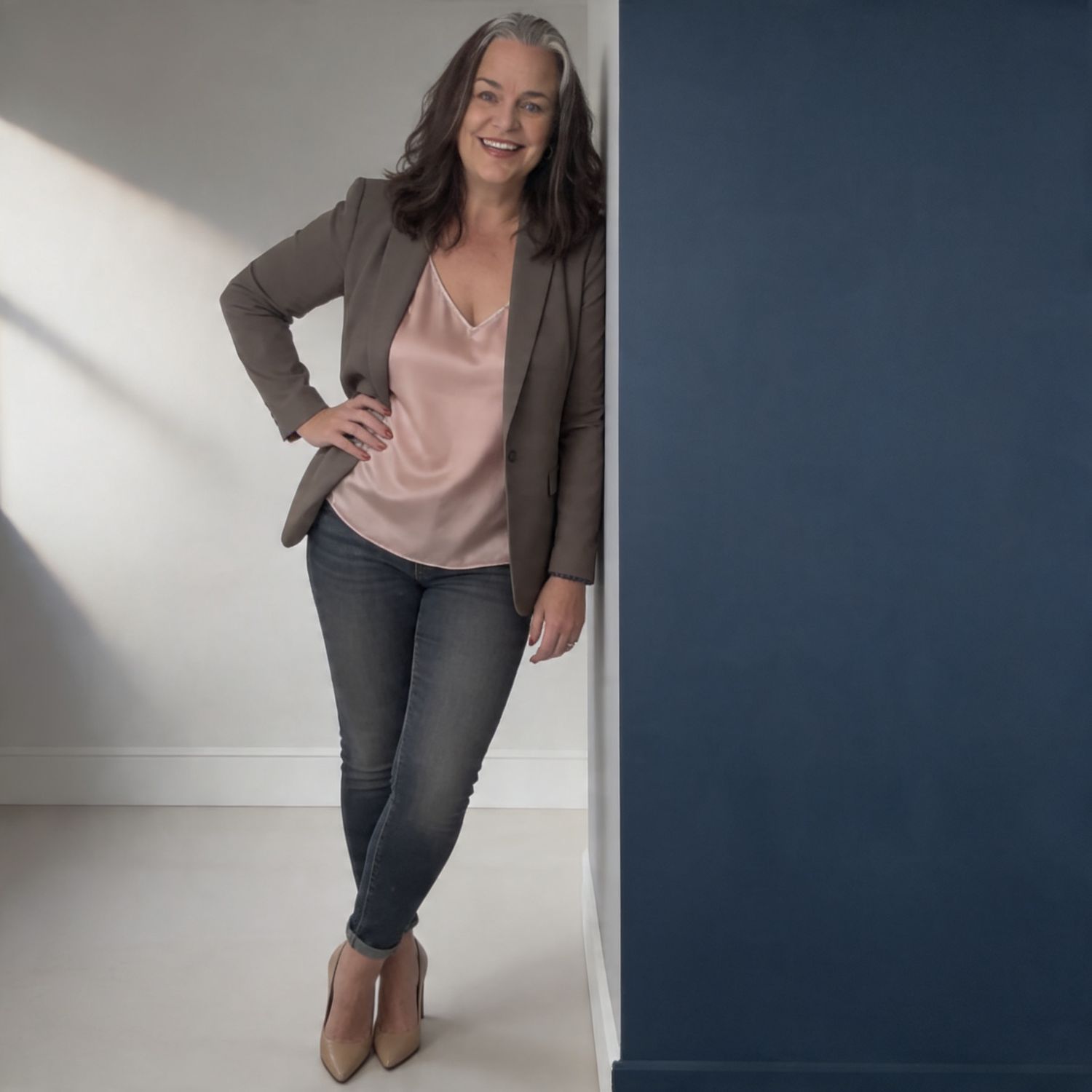 Smiling female photographer in brown blazer and jeans leaning against a dark blue wall.