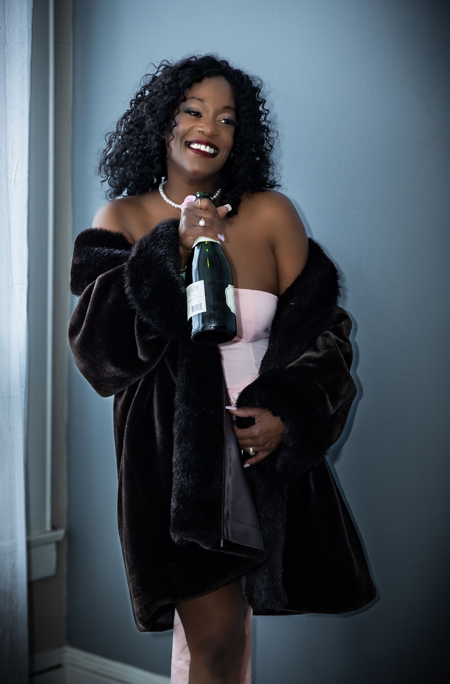 A smiling woman in a pink dress and dark fur coat holds a champagne bottle, wearing a pearl necklace.