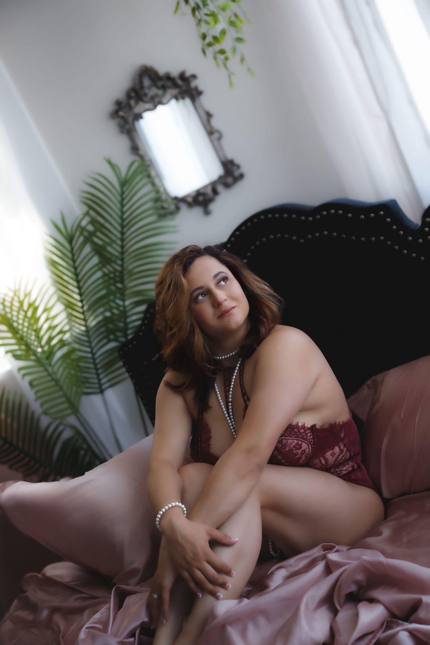 Woman in burgundy lace lingerie and pearl necklaces sits on a bed with pink satin sheets, gazing up in a boudoir portrait.