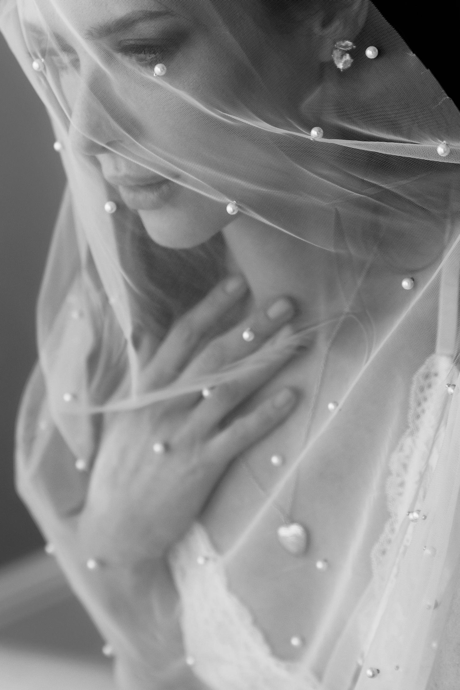 Black and white bridal portrait of a bride draped in a pearl-embellished veil, hands clasped gracefully at her chest.