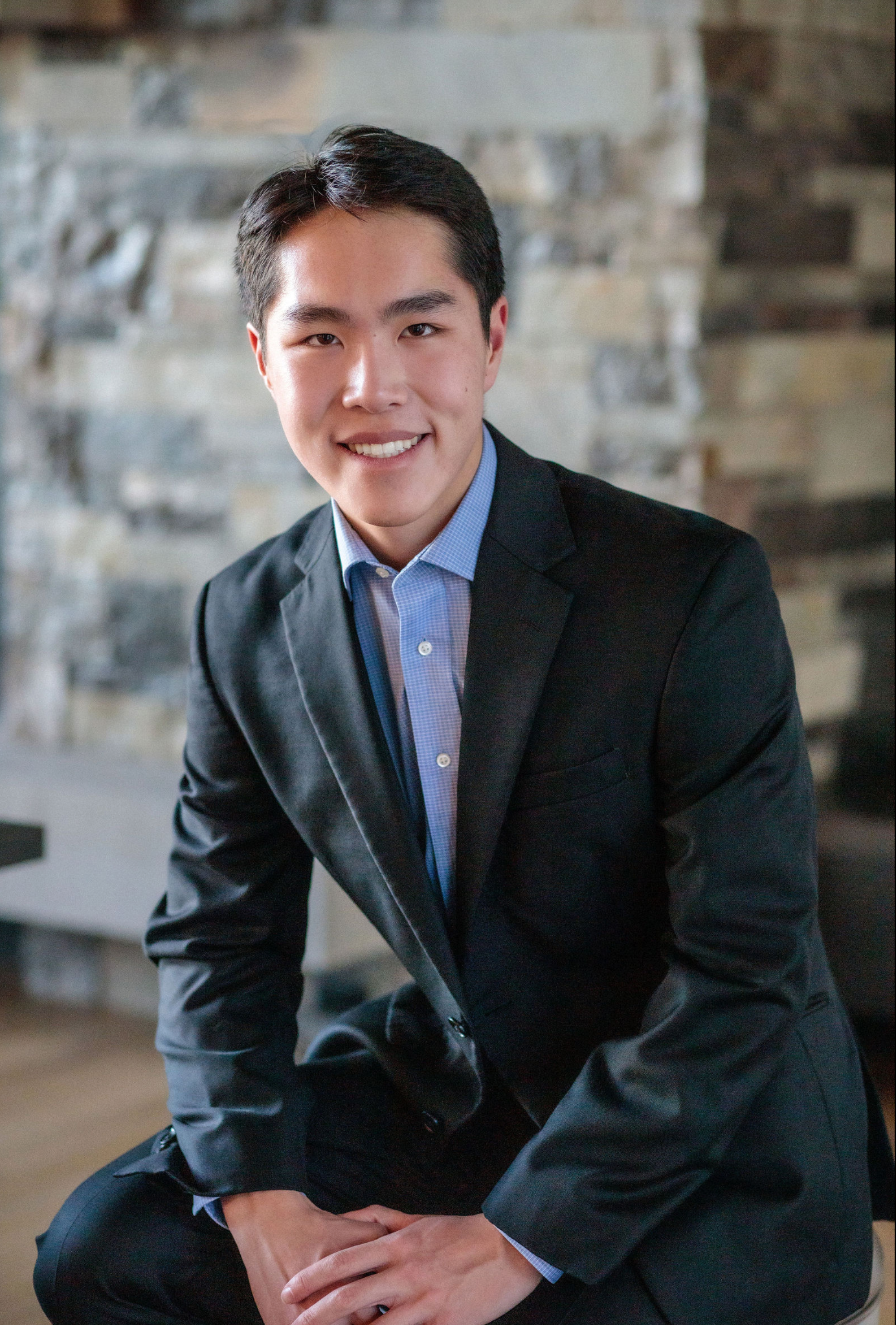 Young professional man in black suit and light blue shirt smiling confidently in a modern stone-accented interior.