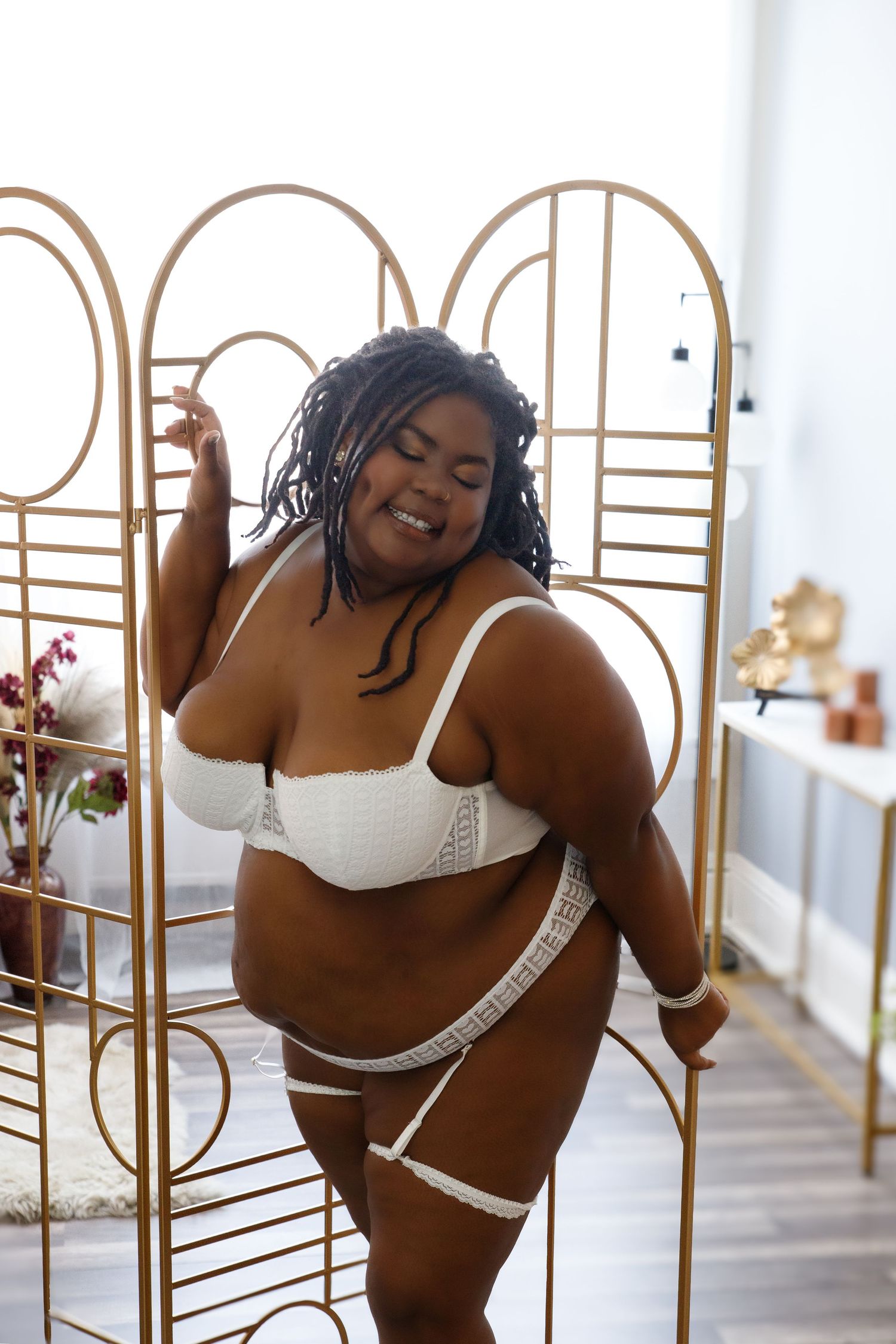 Plus-size woman posing confidently in white lace lingerie set beside a gold decorative room divider.