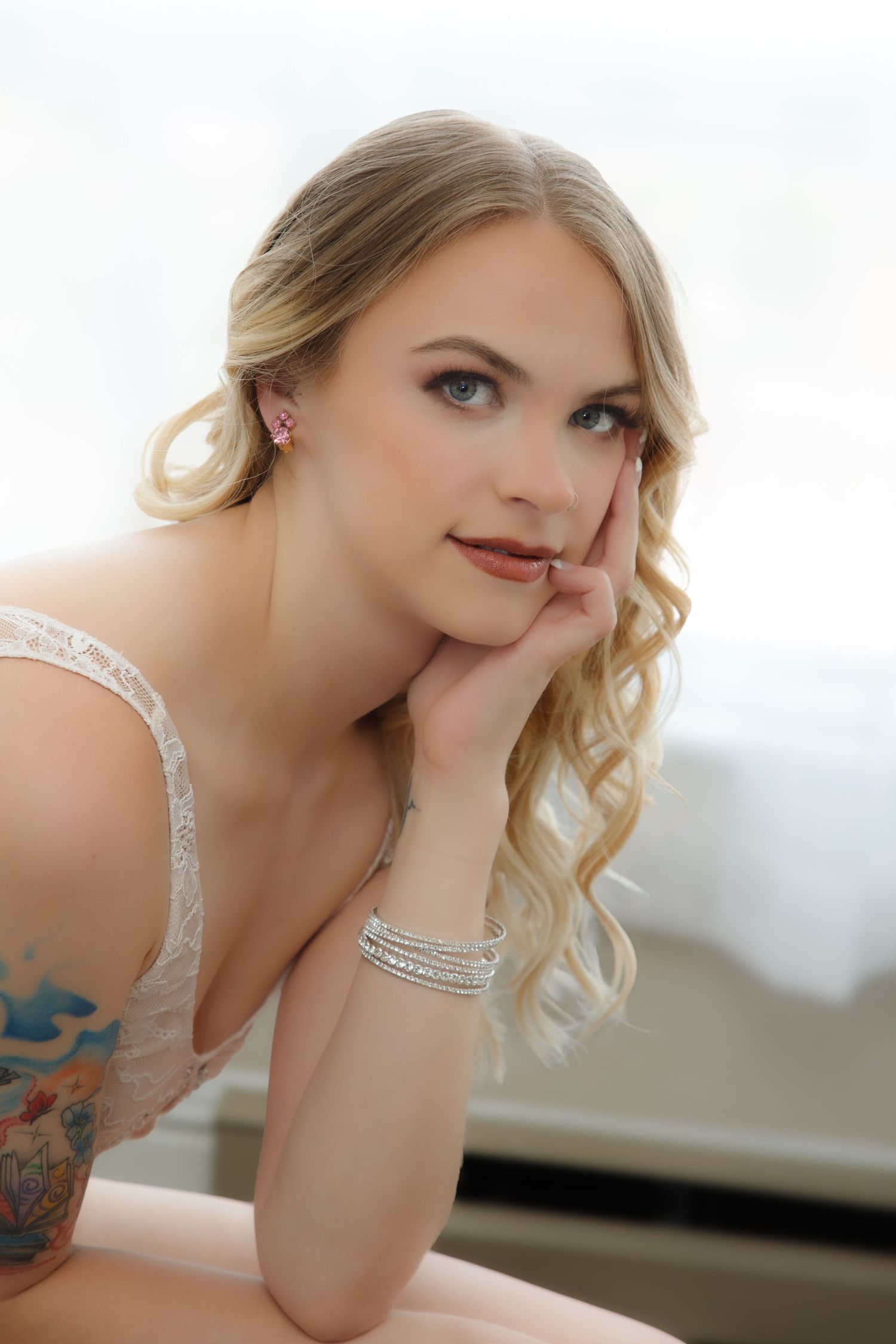 Blonde woman with blue eyes, silver bracelet, pink earrings, and tattoo poses thoughtfully with hand on cheek.