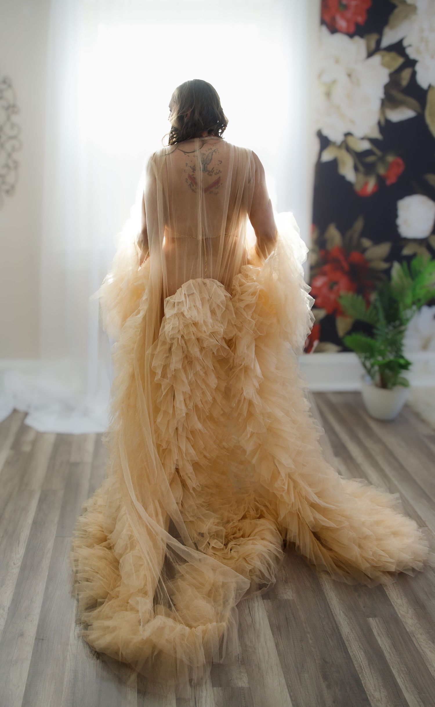 Woman in sheer champagne tulle gown with ruffled train, viewed from behind near bright window.