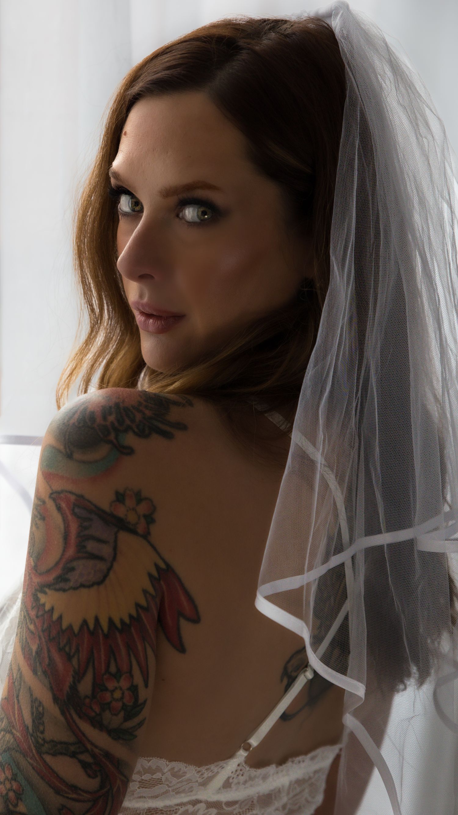 A tattooed bride wearing a white veil and lingerie looks over her shoulder, showcasing colorful arm tattoos.