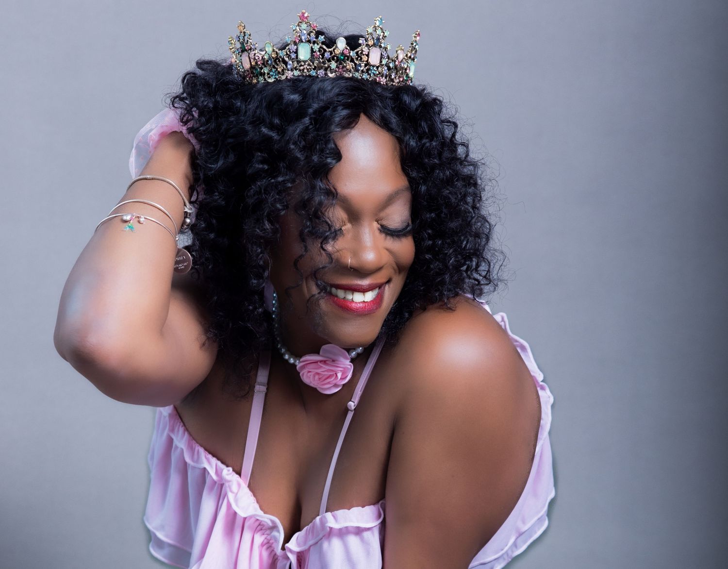 Smiling Black woman wearing a decorative crown and pink outfit, posing confidently with hand in curly hair.
