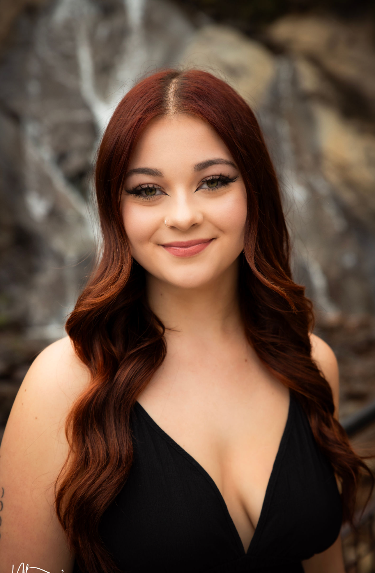 Young woman with long auburn hair wearing a black dress smiling outdoors near a waterfall.