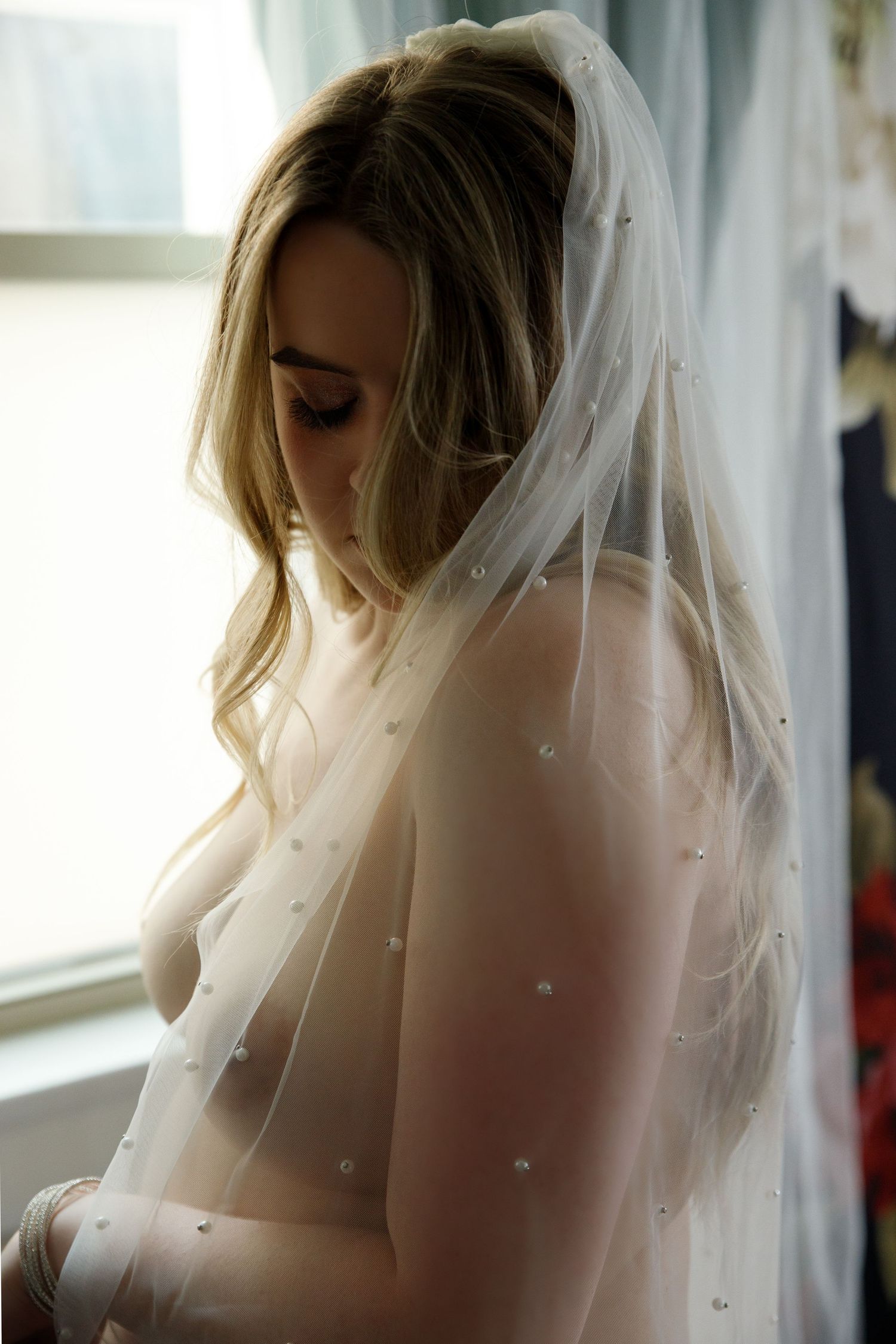 Bride wearing a pearl-embellished white veil near a window in soft natural light.