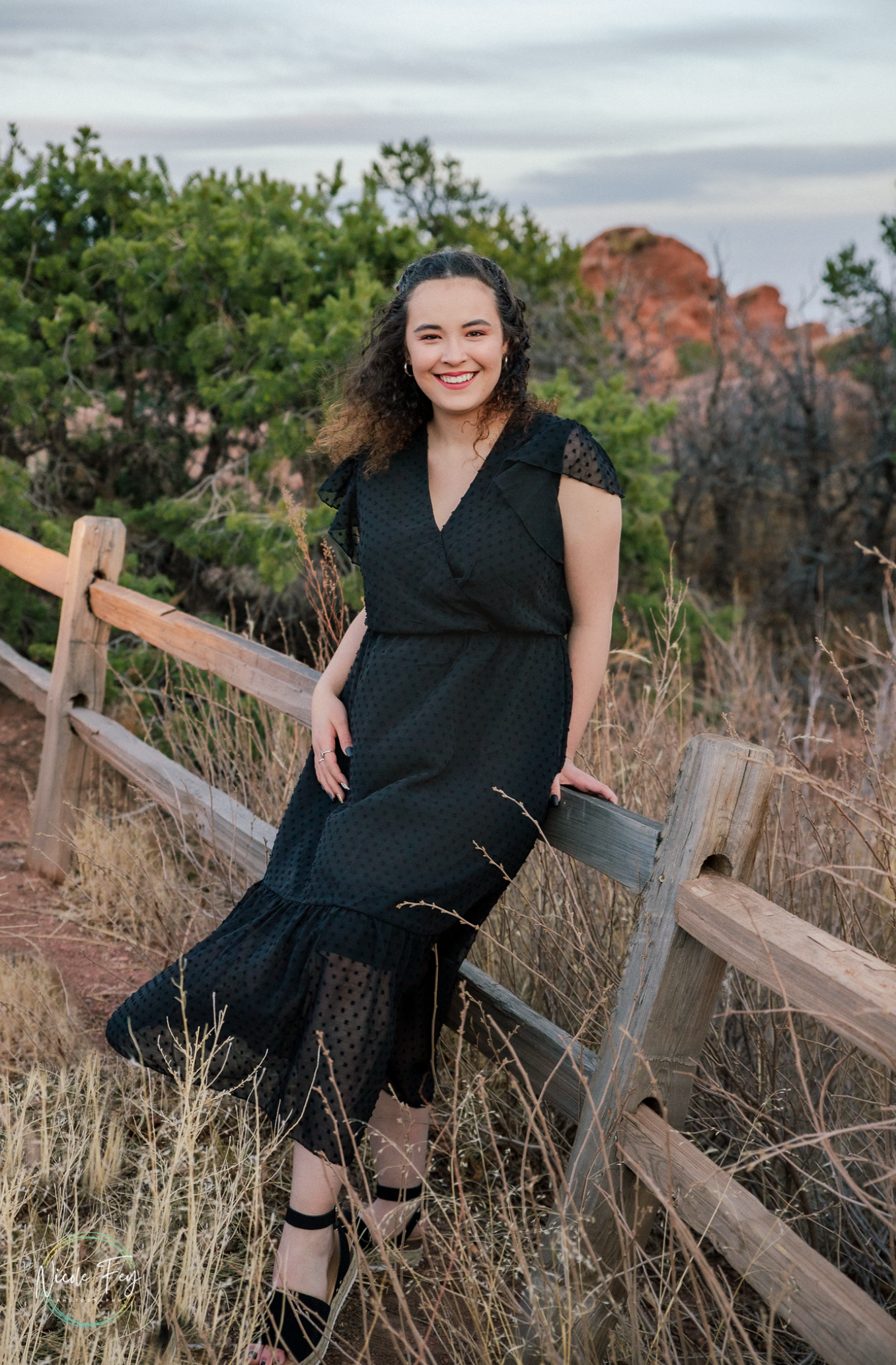A smiling young woman in a black lace dress poses against a wooden fence in a scenic desert landscape.