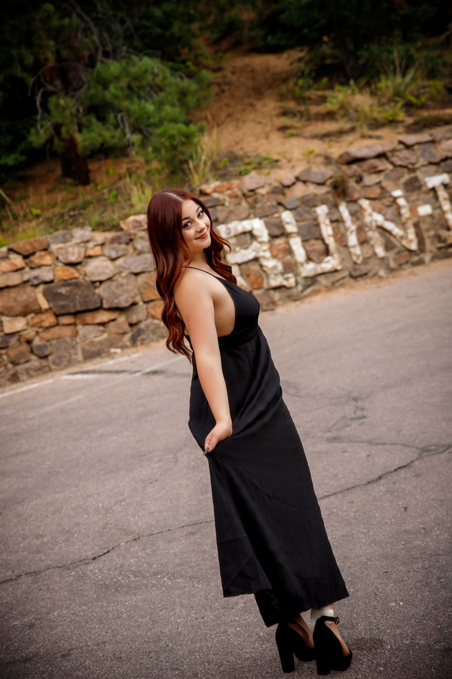 Woman in black maxi dress and heels posing on a road near a stone wall with letters carved into it.