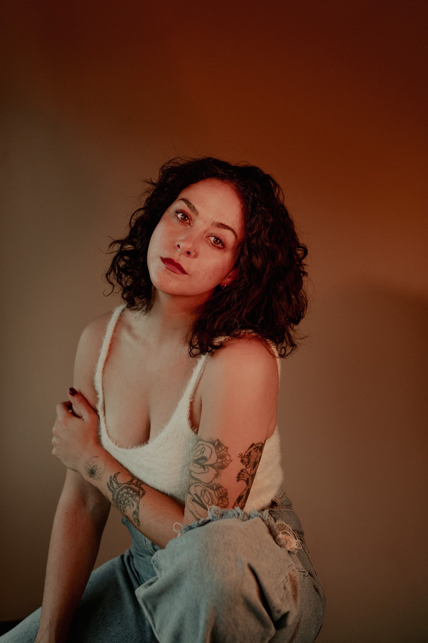 A tattooed woman with curly dark hair poses in a white top and jeans under warm amber studio lighting. Photographer for artists in Colorado.