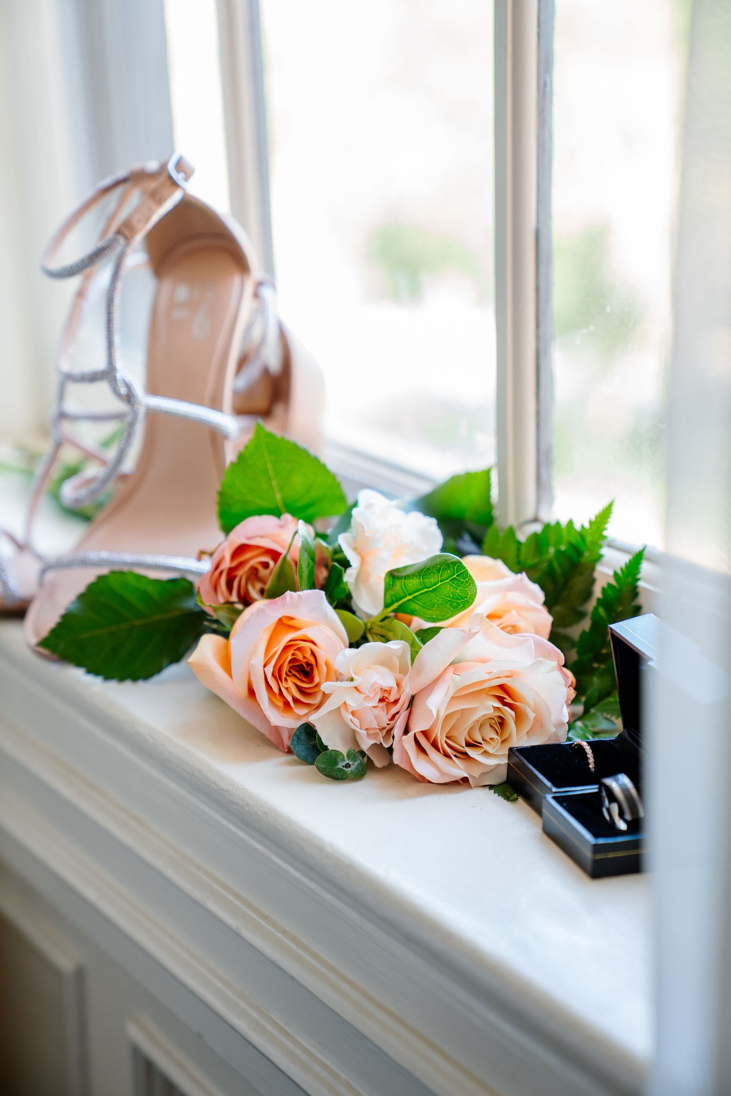 A bridal flat lay featuring blush roses, a ring box, and strappy heeled shoes on a windowsill. Photography of wedding day details in Denver.