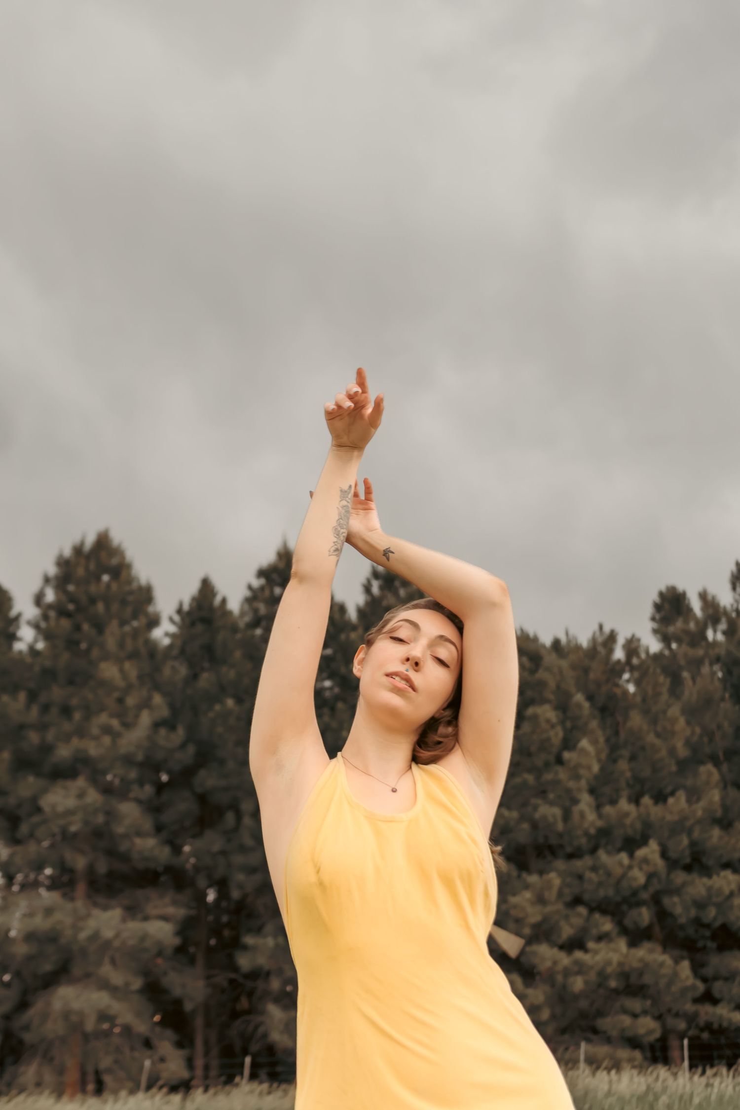 Woman in yellow dress posing with arms raised against cloudy sky and pine trees backdrop.