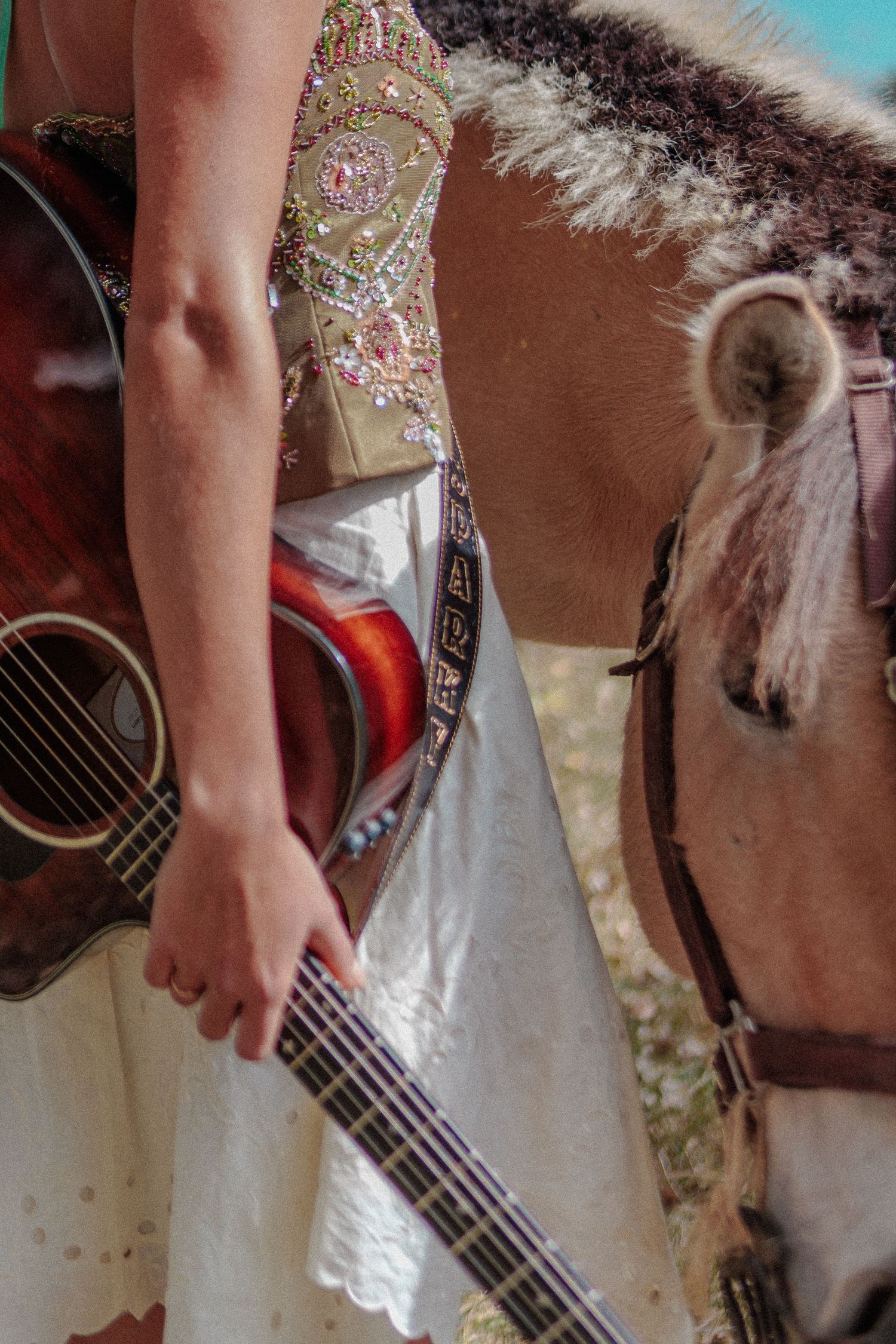 Woman in embroidered dress holds acoustic guitar beside a horse in outdoor setting. Local Colorado musician.