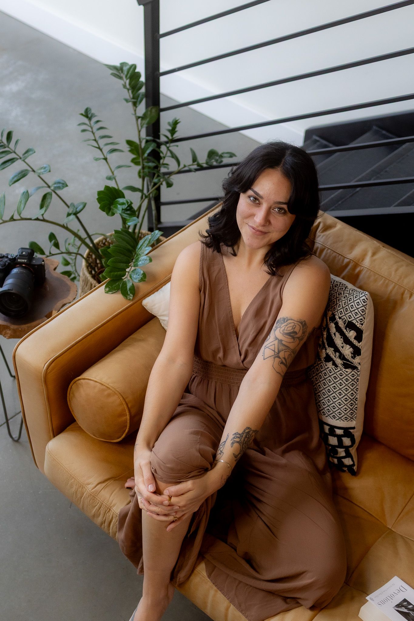 Brunette woman with arm tattoo in brown satin dress sitting on leather armchair in modern interior.