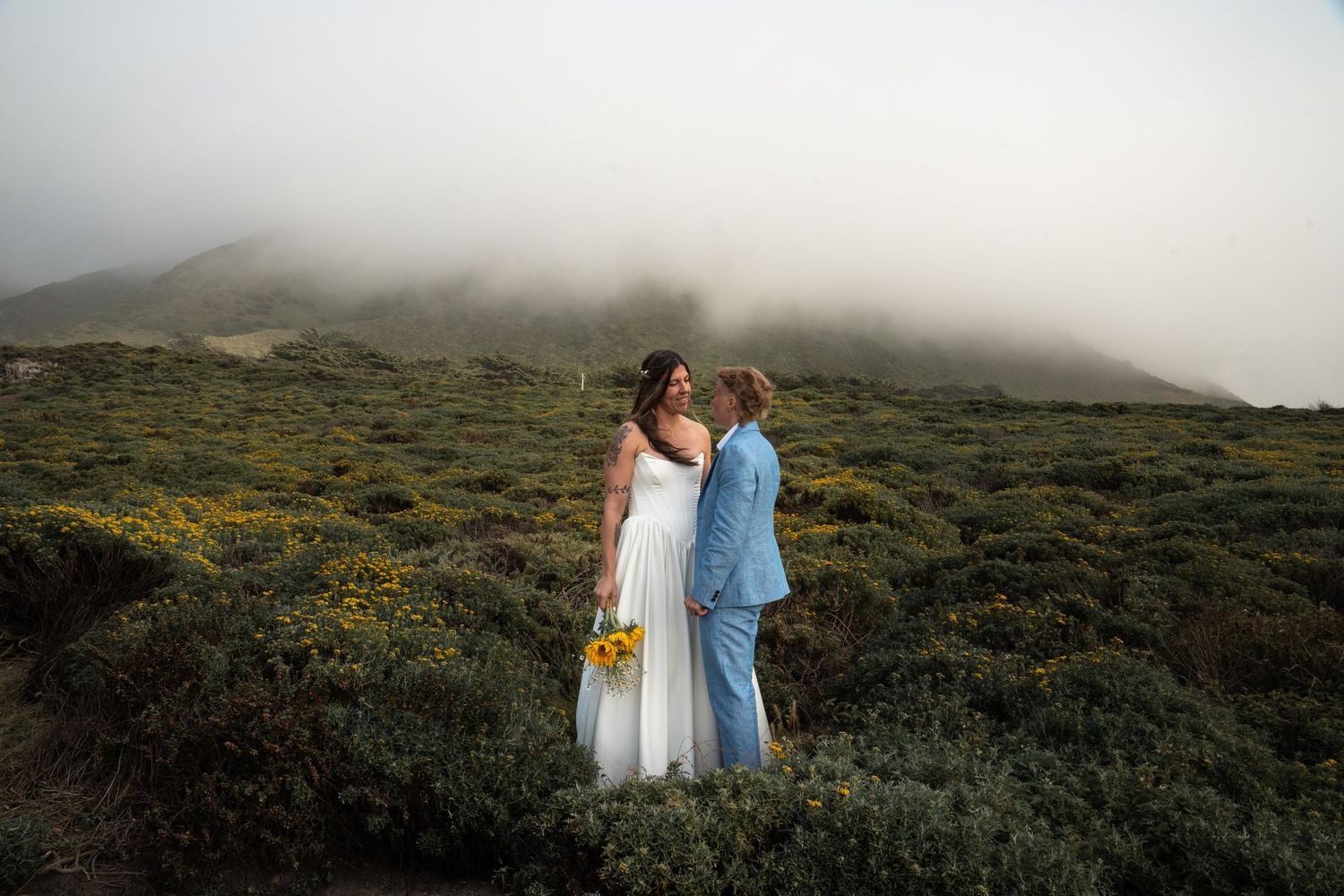 Same-sex wedding couple sharing a kiss surrounded by foggy green coastal landscape.