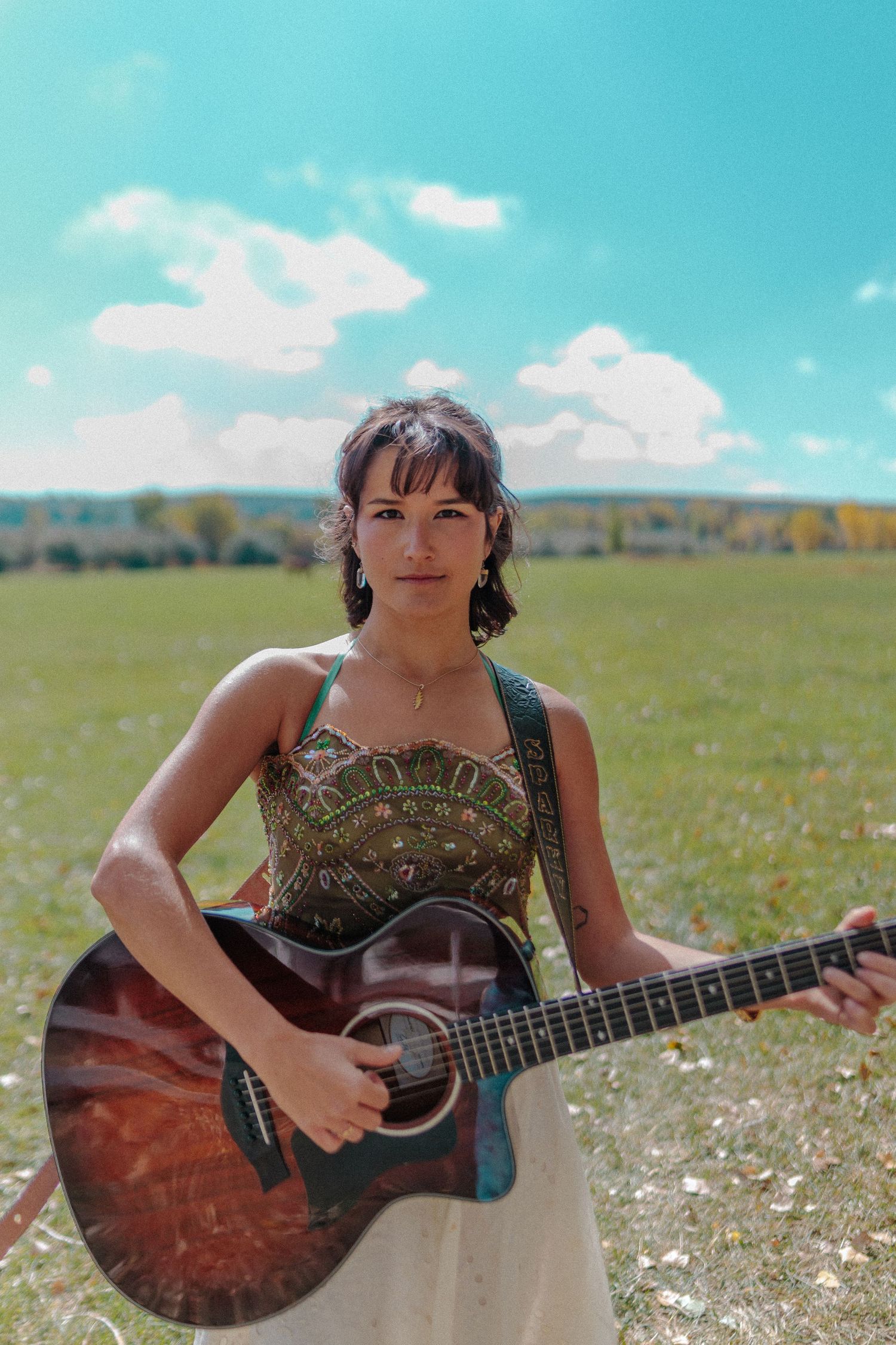 Young woman in embroidered top holds acoustic guitar in a sunny green field. Photos for Denver musician.