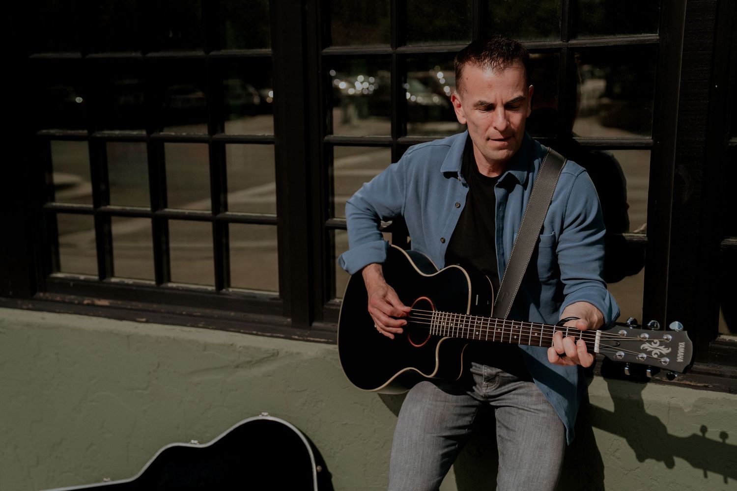 Man playing acoustic guitar outdoors, seated casually in jeans and denim shirt with guitar case nearby.