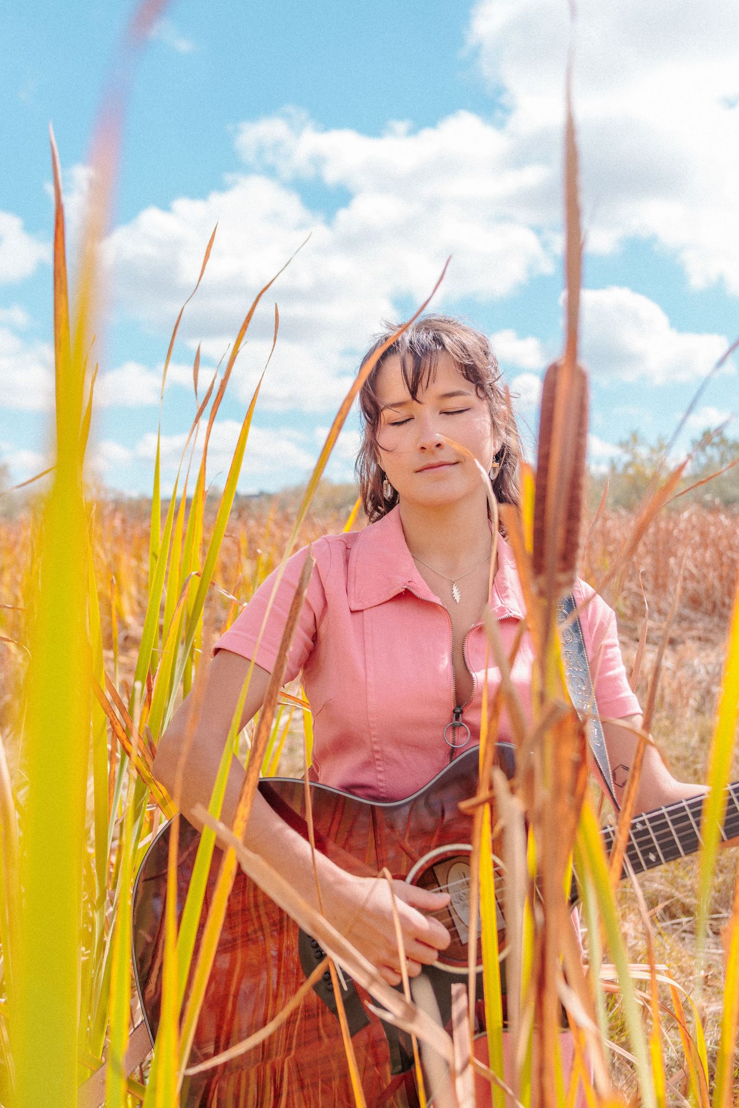 Young woman with closed eyes holding a guitar among tall cattail reeds in a sunny field. Photography for local Colorado musician.