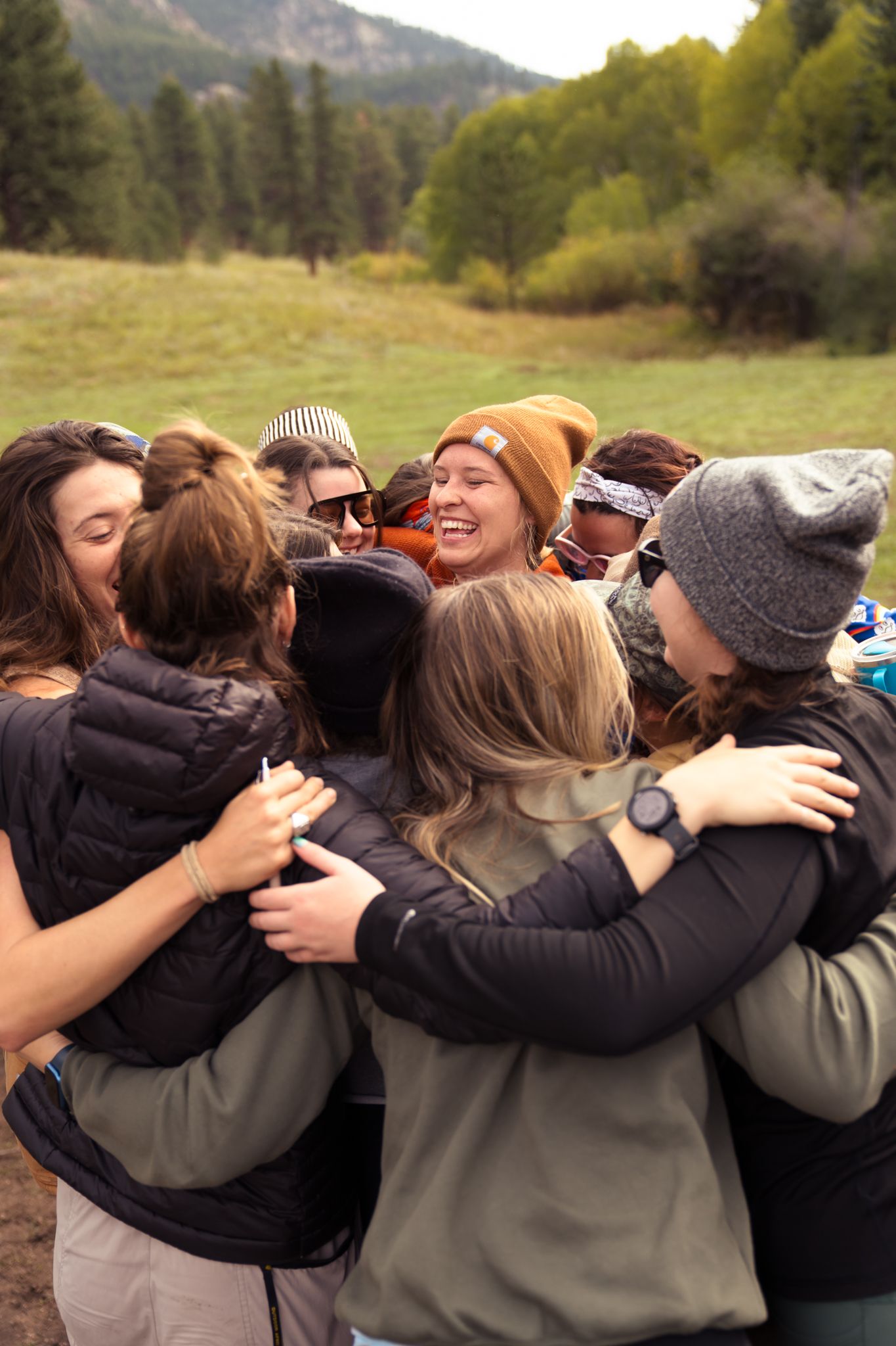 Group of women in outdoor gear sharing a warm group hug in a mountain meadow surrounded by pine trees.