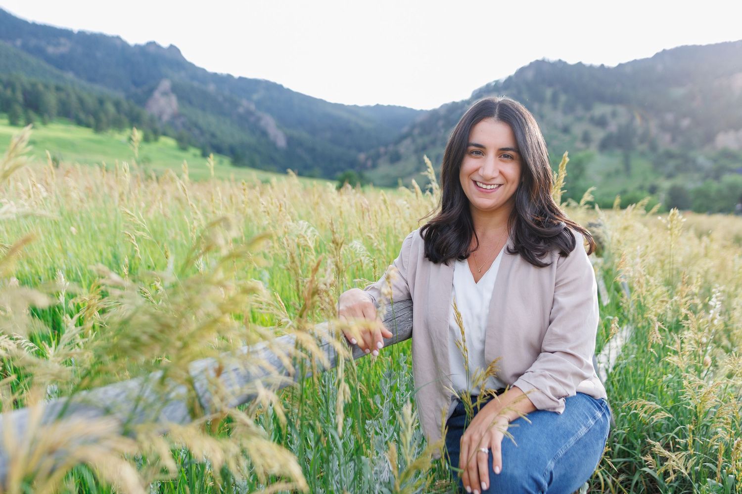 A smiling woman crouches in a lush green field of tall grass with mountain scenery behind her. Headshot photography for Colorado therapist.
