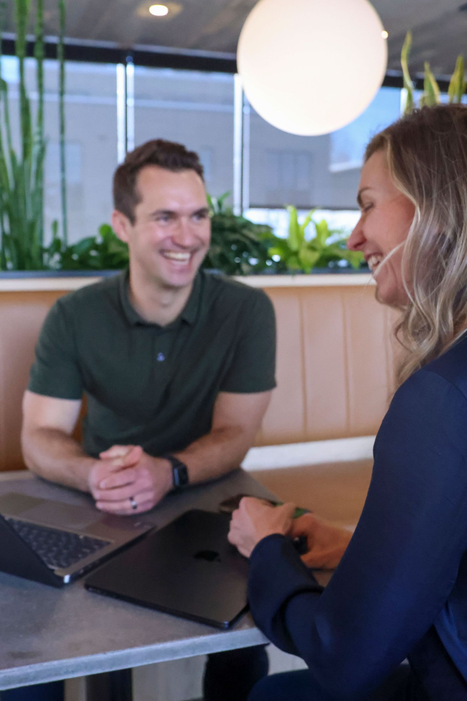 Man and woman smiling and laughing together at cafe table with laptop in modern office setting.