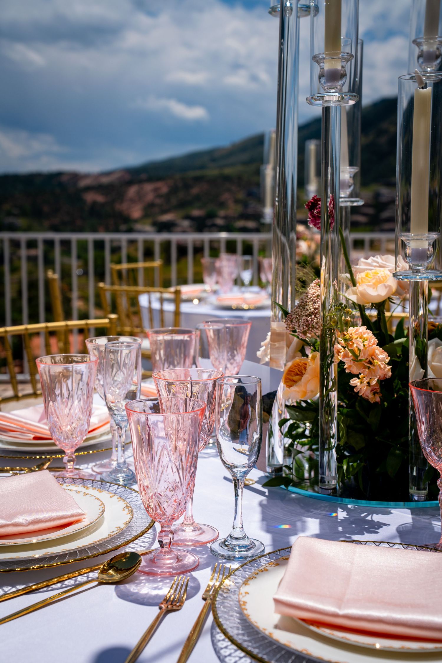 An elegant outdoor wedding reception table set with pink crystal glasses, gold cutlery, and floral centerpieces. Photography in Morrison, CO