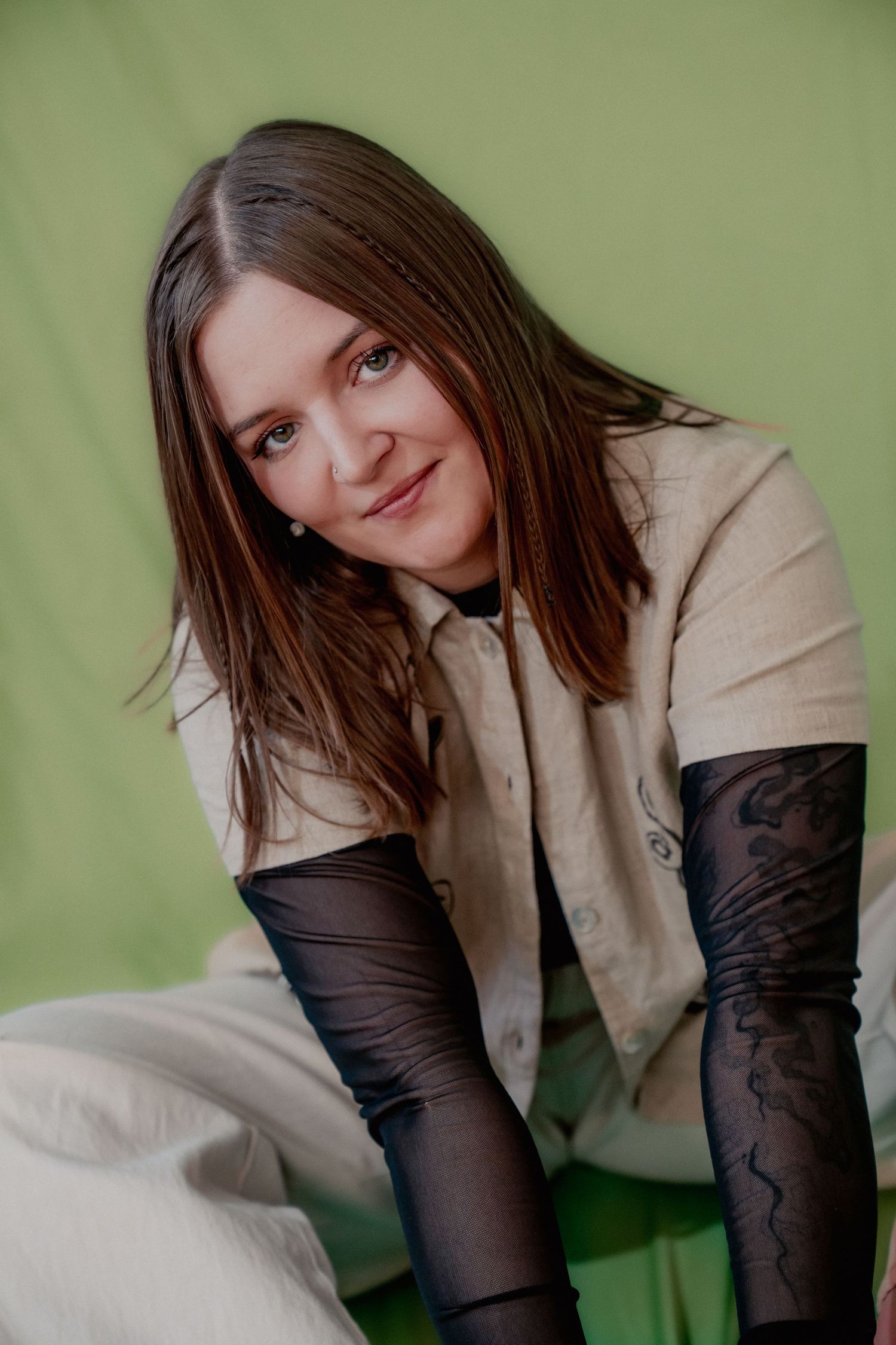 Woman with brown hair wearing a beige shirt and black lace sleeves posing against a green background. Denver musician.