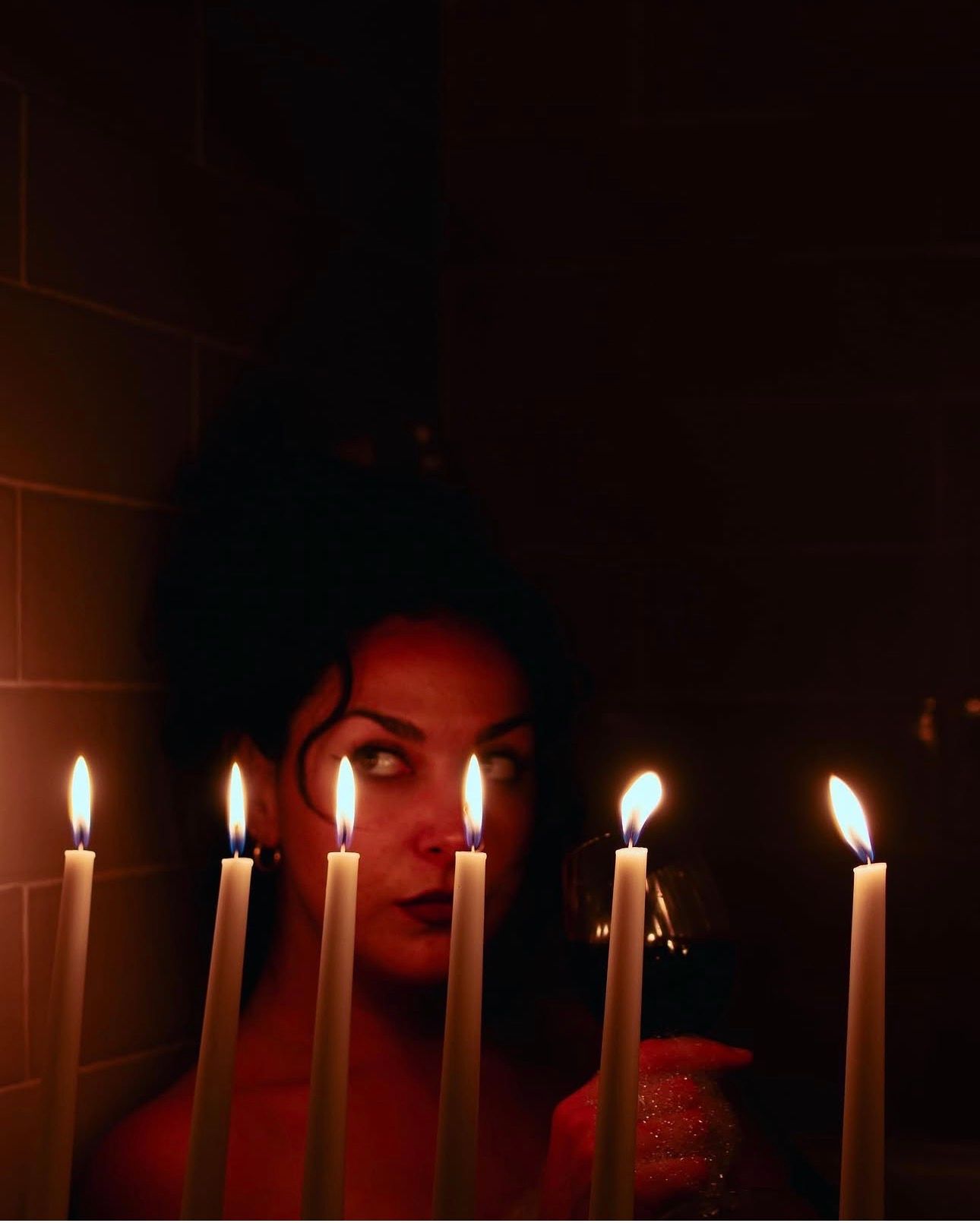 Woman gazing mysteriously behind five lit white candles in a dark, moody setting.