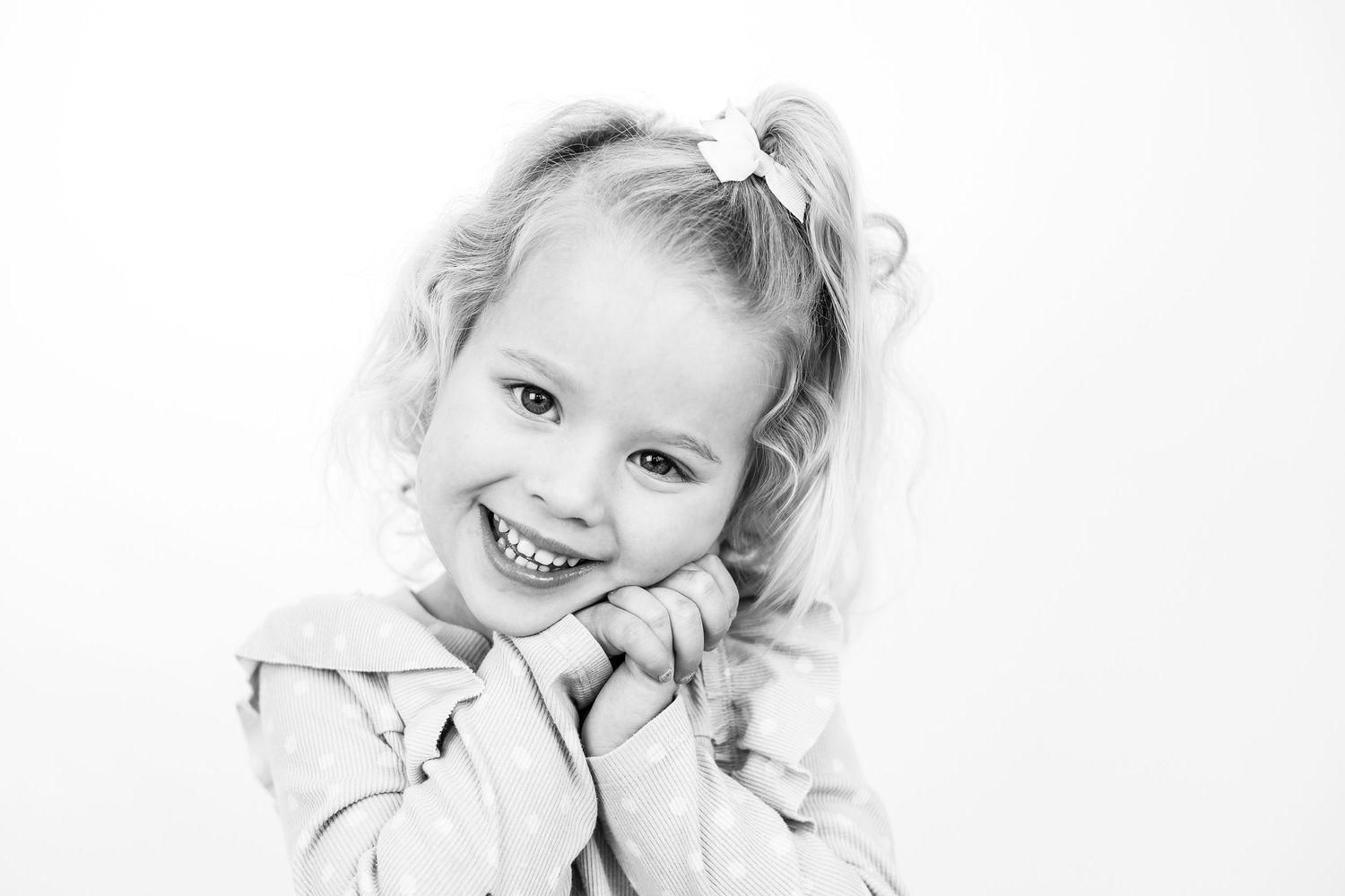 Black and white portrait of a young curly-haired girl smiling brightly with hands clasped under her chin.