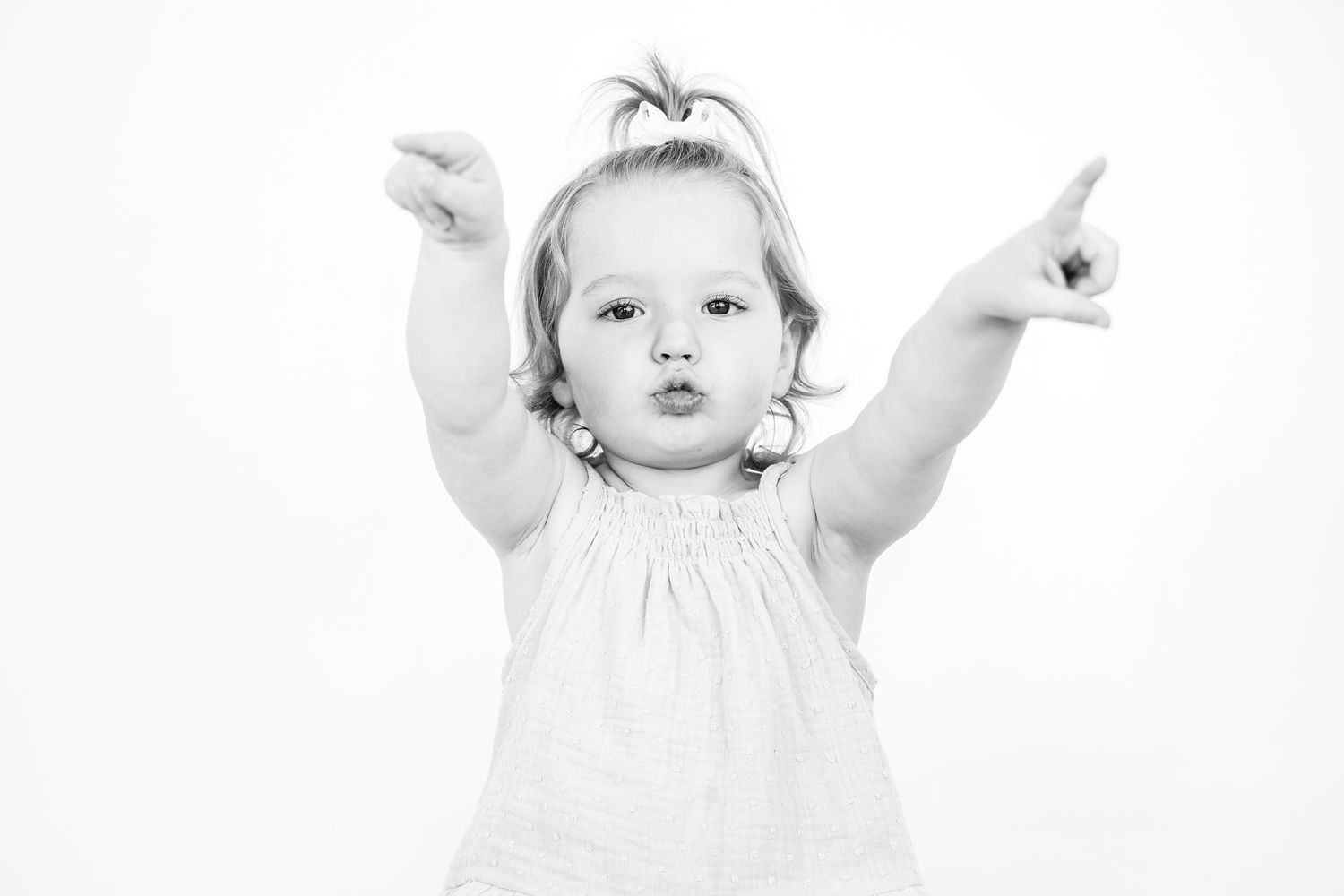 Black and white photo of a toddler in a white dress pointing both arms upward with puckered lips.