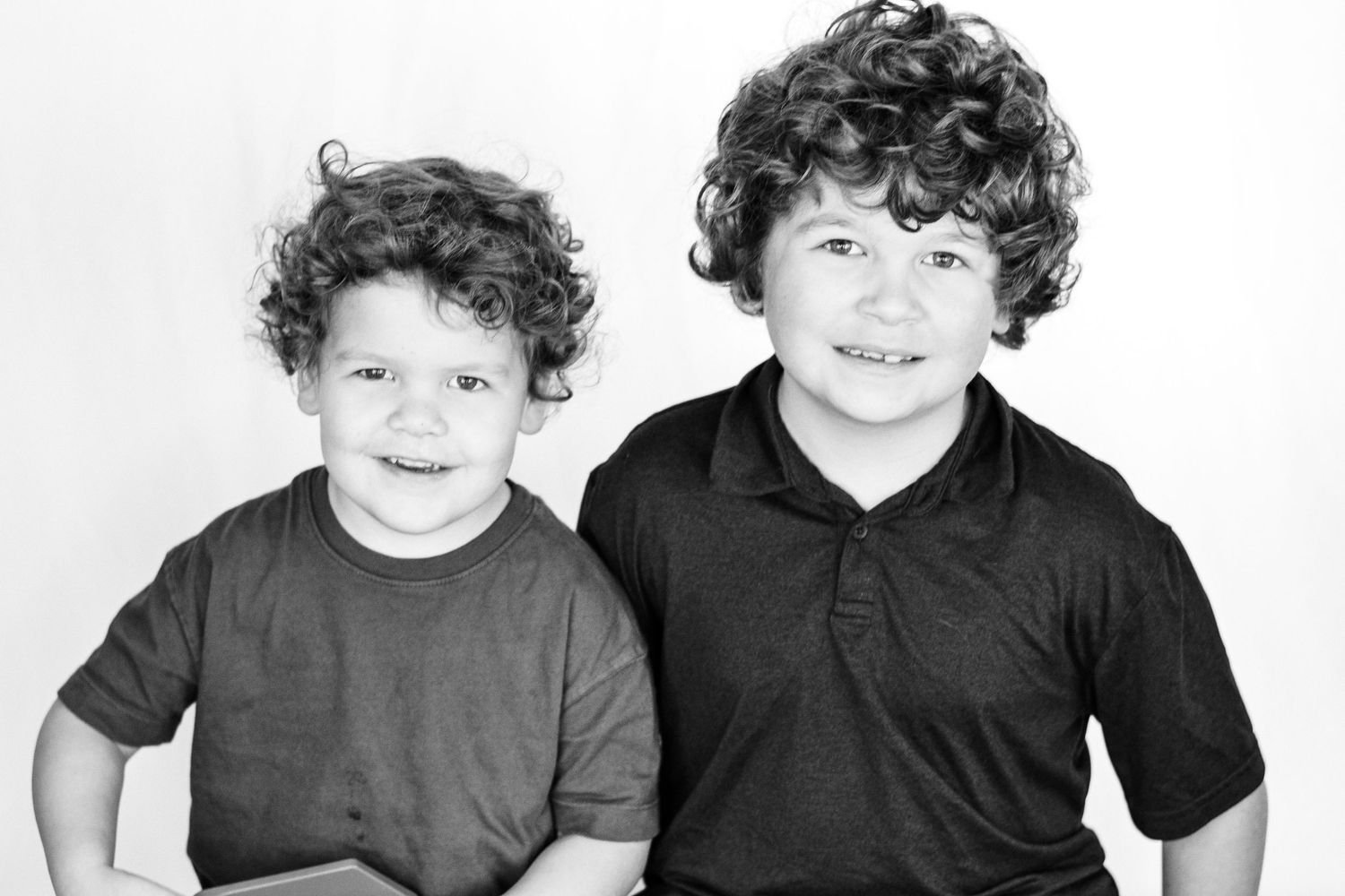 Two curly-haired brothers smile together in a charming black and white studio portrait.