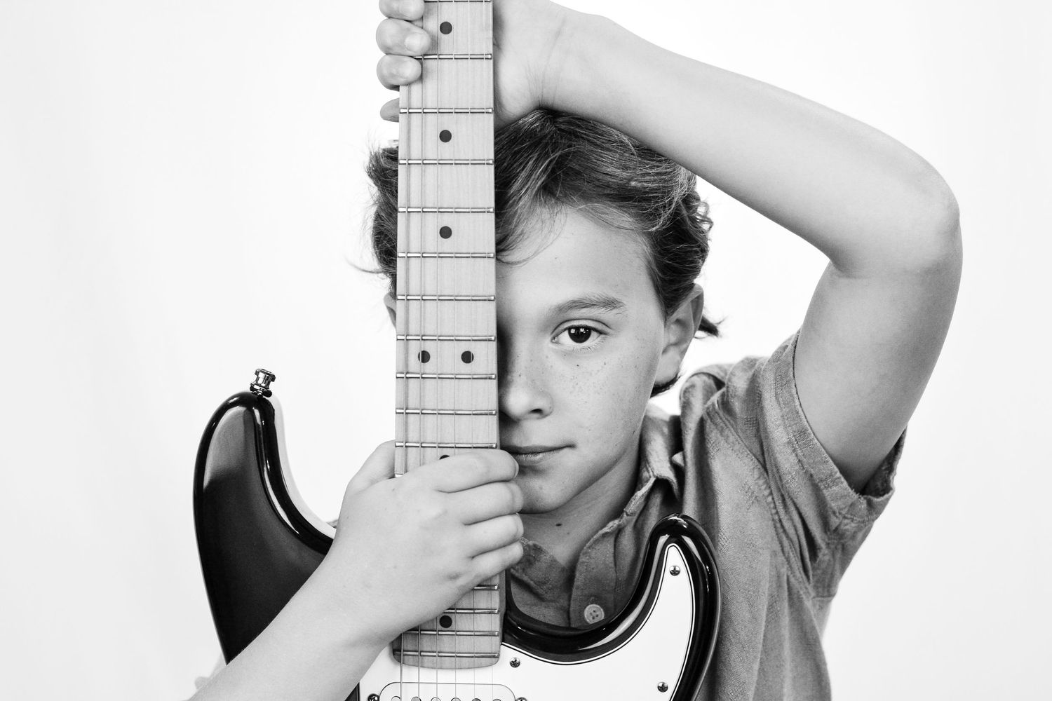 Black and white photo of a young child peeking out from behind an electric guitar held vertically.