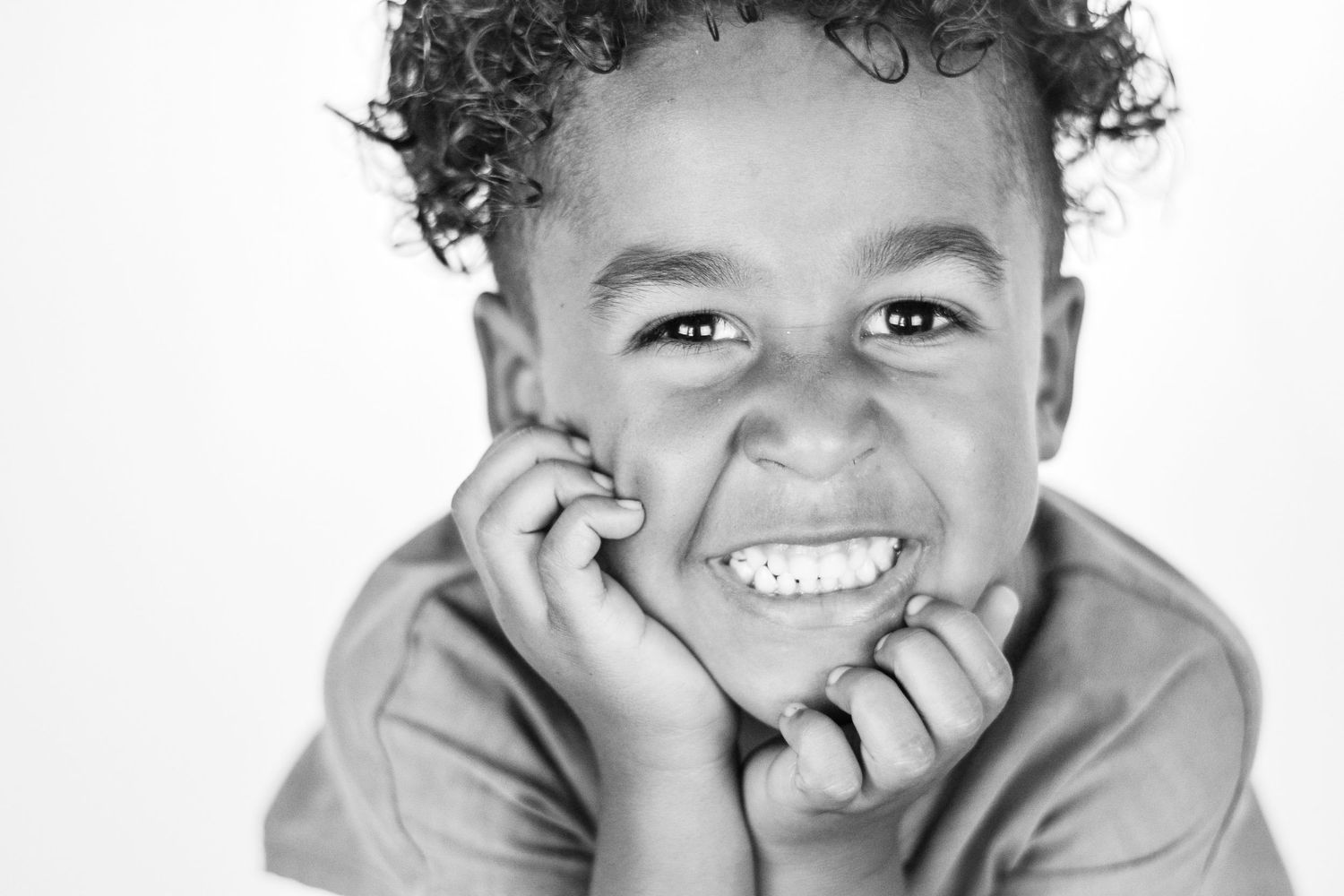 Black and white portrait of a young curly-haired boy grinning widely with hands pressed against his cheeks.