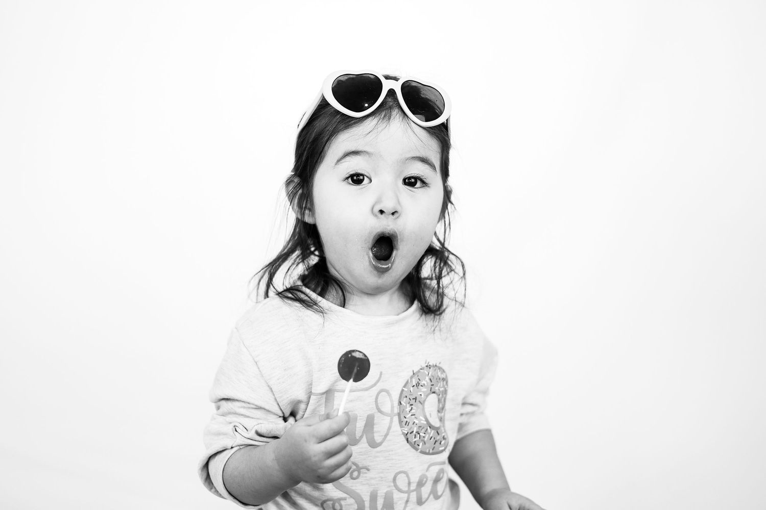 Black and white photo of a young girl with sunglasses on her head, holding a lollipop with mouth wide open.